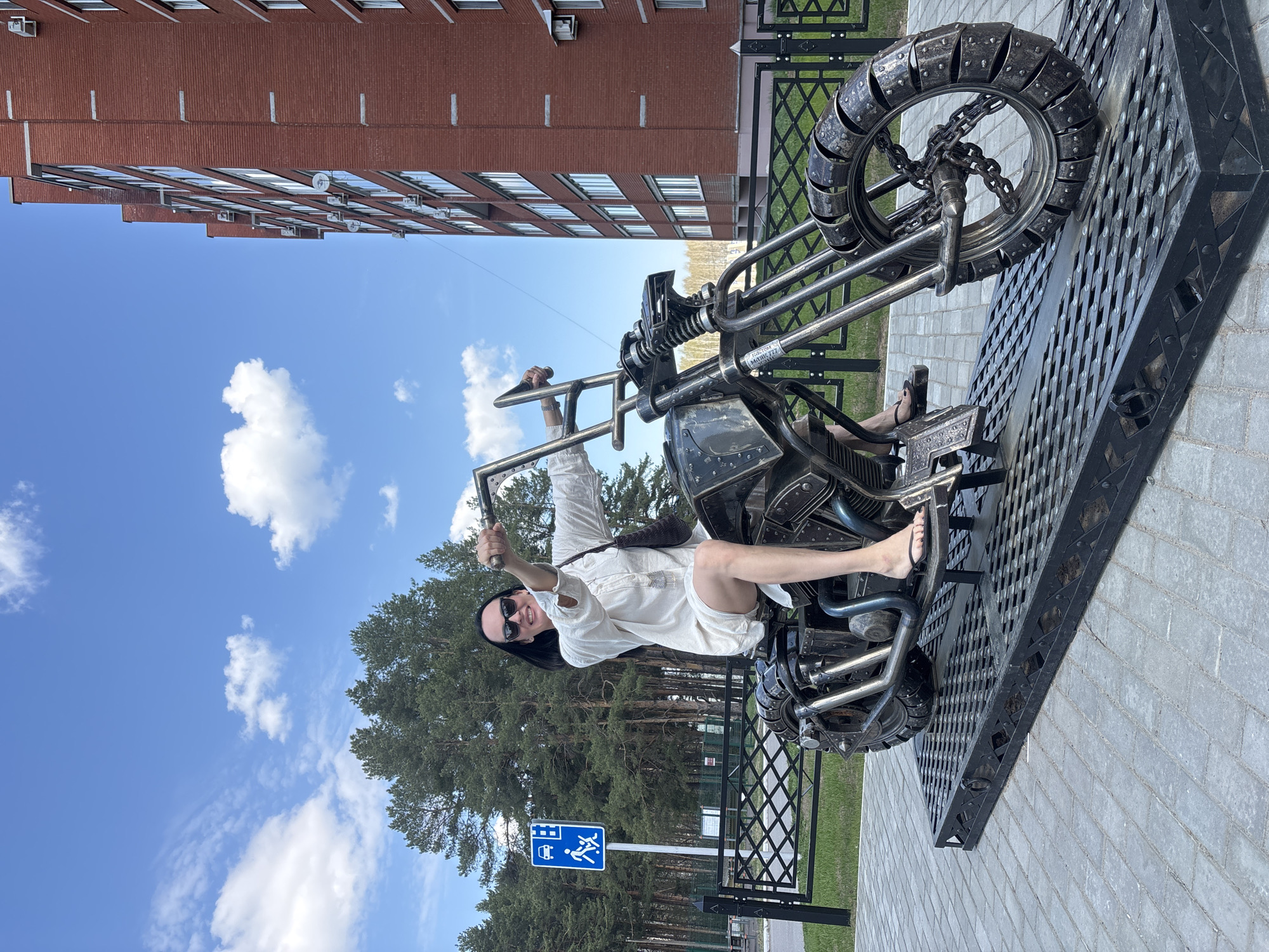 A woman in a white dress, black sunglasses, and black sandals is sitting on a metal motorcycle sculpture in Chaikovsky, Russia. She is holding up one hand in a thumbs-up gesture. The sculpture sits on a metal platform on a paved surface, with a metal fence and a bus stop sign visible in the background. Behind the fence is a grassy area and a dense pine forest. A multi-story brick building with windows is in the background on the left. The sky is blue with scattered white clouds.