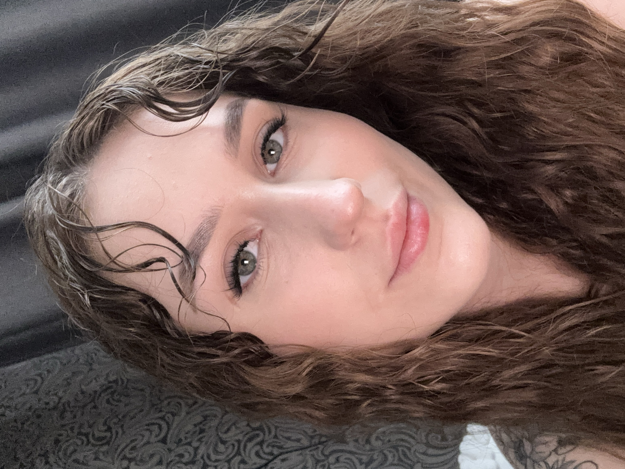 A female individual is depicted in a close-up portrait, facing the camera directly, in Марковский, Russia. Her long, dark brown, wavy hair appears damp, with several strands across her forehead. Facial features include green eyes with visible eyelashes, defined eyebrows, and lips gently curved into a slight smile. A small blemish is present on her upper right cheek. On the subject's left shoulder, a tattoo featuring an elaborate floral or scroll-like design is partially visible. The immediate background to her left features a dark grey patterned textile with a swirling motif, while the background to her right is uniformly dark grey or black.
