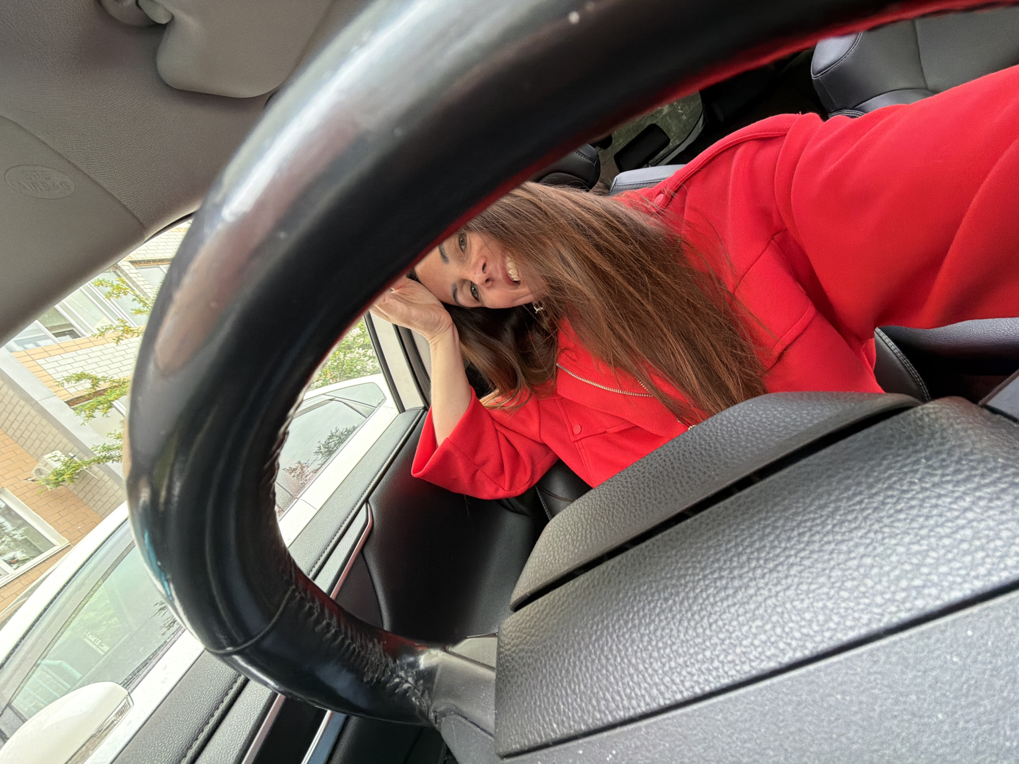 A woman with long brown hair wearing a red jacket is inside a car. She is smiling and leaning her head on her hand near the car window. The car's steering wheel is prominently visible in the foreground. Outside the car window, a building is visible, along with parked cars and trees. There is no way to ascertain if the photo was taken in Birobidzhan, Russia.
