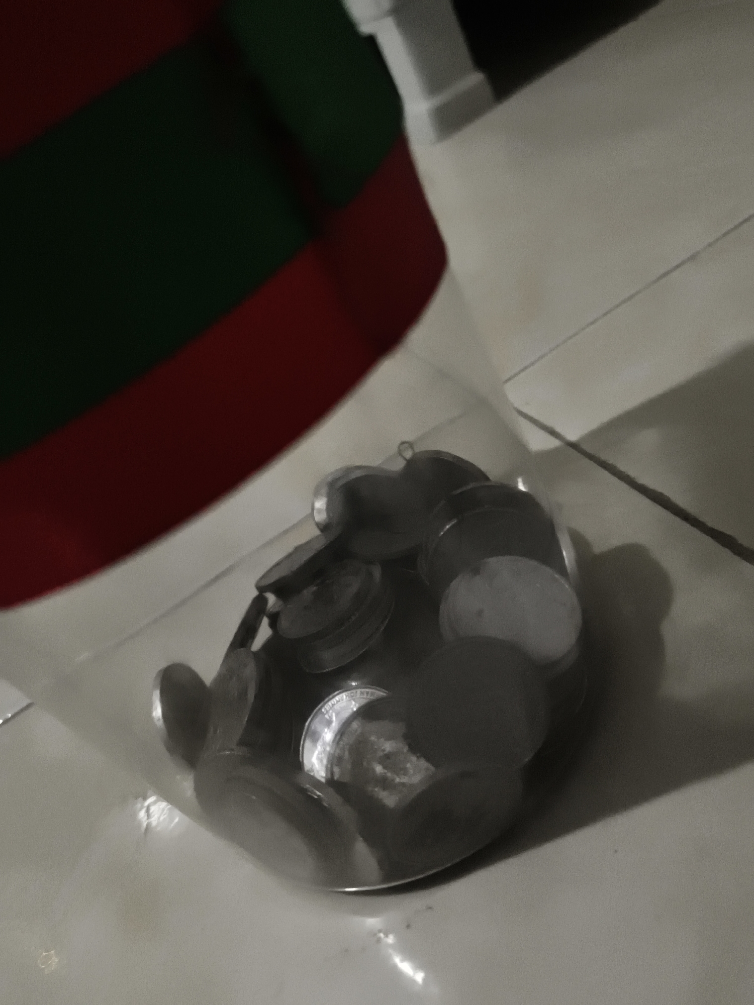 The image shows a clear plastic bag filled with coins. The bag is partially obscured by a red and green striped object, possibly a piece of fabric or clothing, positioned in the upper left quadrant of the image. The coins are metallic and appear to be of various denominations. They are piled inside the bag, creating a dense collection. The background consists of a light-colored tiled surface, likely a floor. The lighting is dim, casting shadows on the tiled surface, suggesting an indoor environment. The scene does not provide any specific geographical markers to indicate a location in Petiken, Indonesia.
