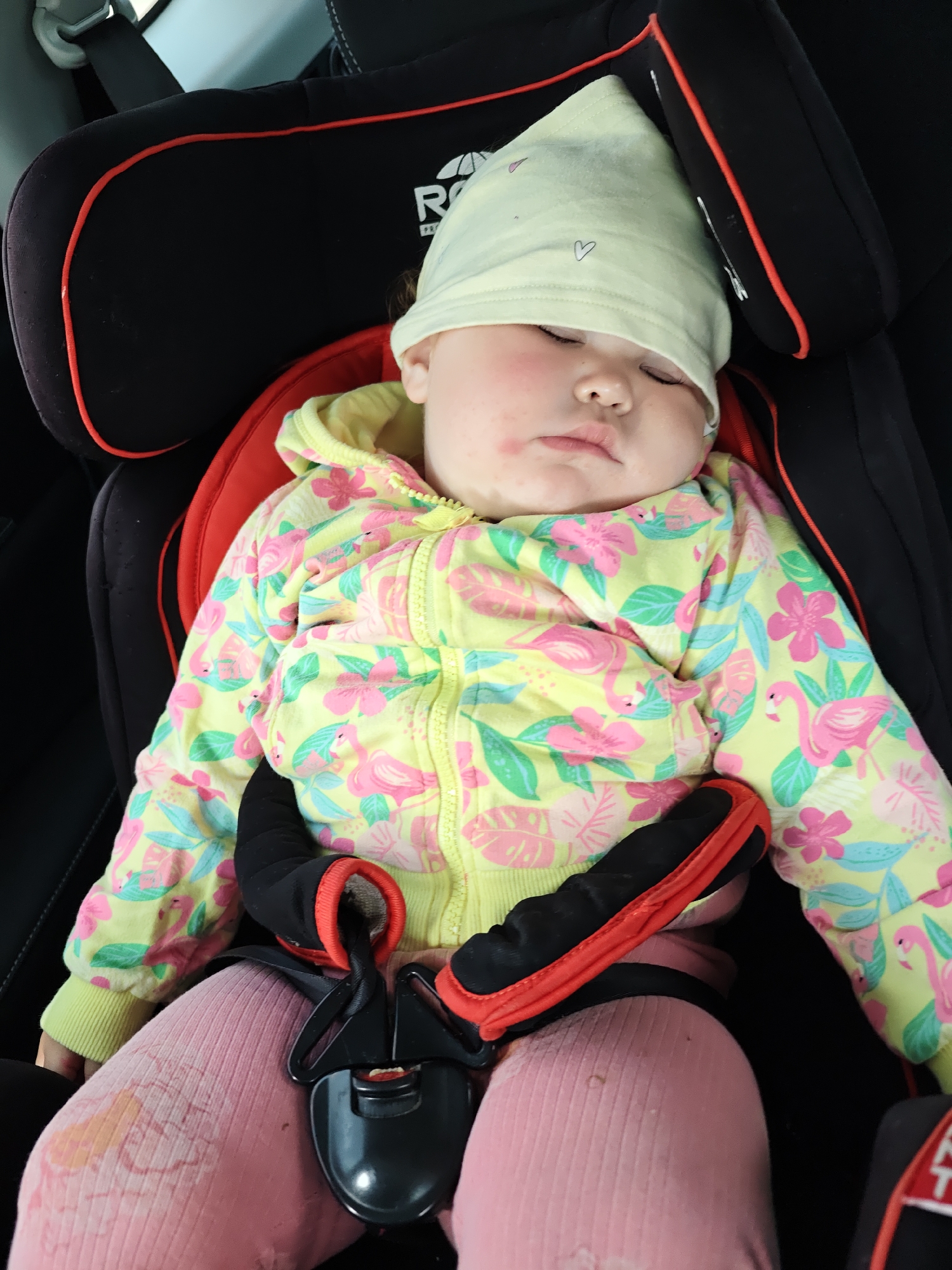 A baby is sleeping in a car seat. The baby is wearing a light yellow beanie, a yellow zip-up hoodie with a pink flamingo and floral pattern, and pink leggings. The car seat is black with red trim, and the baby is secured with a black harness with red padding. The location is assumed, but cannot be determined from the image.