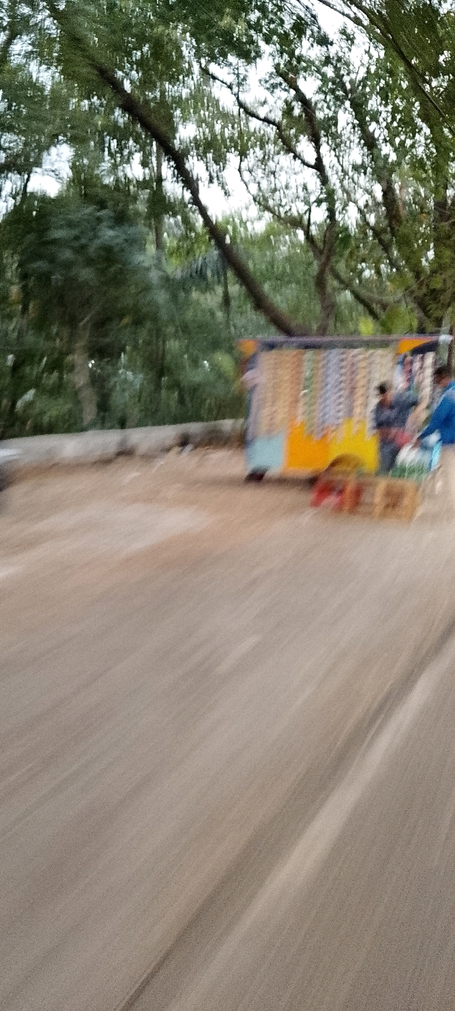 The image shows a blurred outdoor scene with trees in the background and a dirt or gravel surface in the foreground. In the middle ground, there is a mobile stall or cart with colorful items displayed on it. Two figures are partially visible near the stall. One figure is wearing a blue shirt, and another appears to be wearing a darker colored garment. The overall impression is that of a roadside market or vendor stall. The motion blur suggests the image was taken while moving, possibly from a vehicle. The specific location in Depok, Indonesia is not discernible from the image itself, as there are no identifiable landmarks or signs.