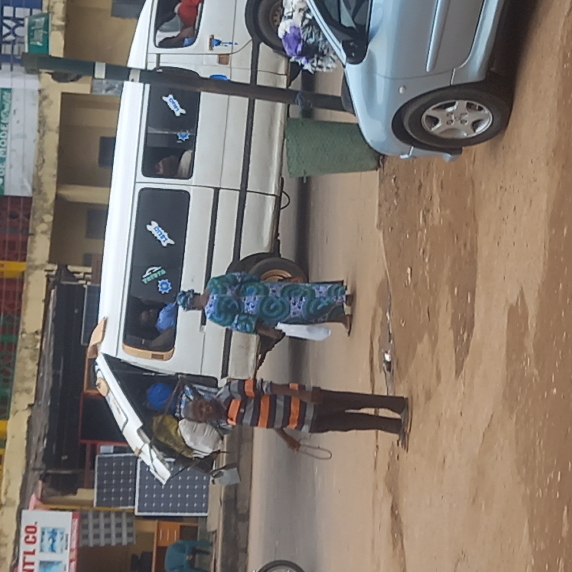 The image depicts an urban scene in Dadi Riba, Nigeria. A white van, partially obscured by a light blue car, is parked on a paved street. A woman in a blue and green patterned dress stands near the van. A man wearing a horizontally striped orange and grey shirt and dark pants holds a wire object in his hand. The surface of the street is reddish-brown. To the left, a building houses what appears to be a shop, with solar panels and other goods visible. Signage on the building is partially obscured.