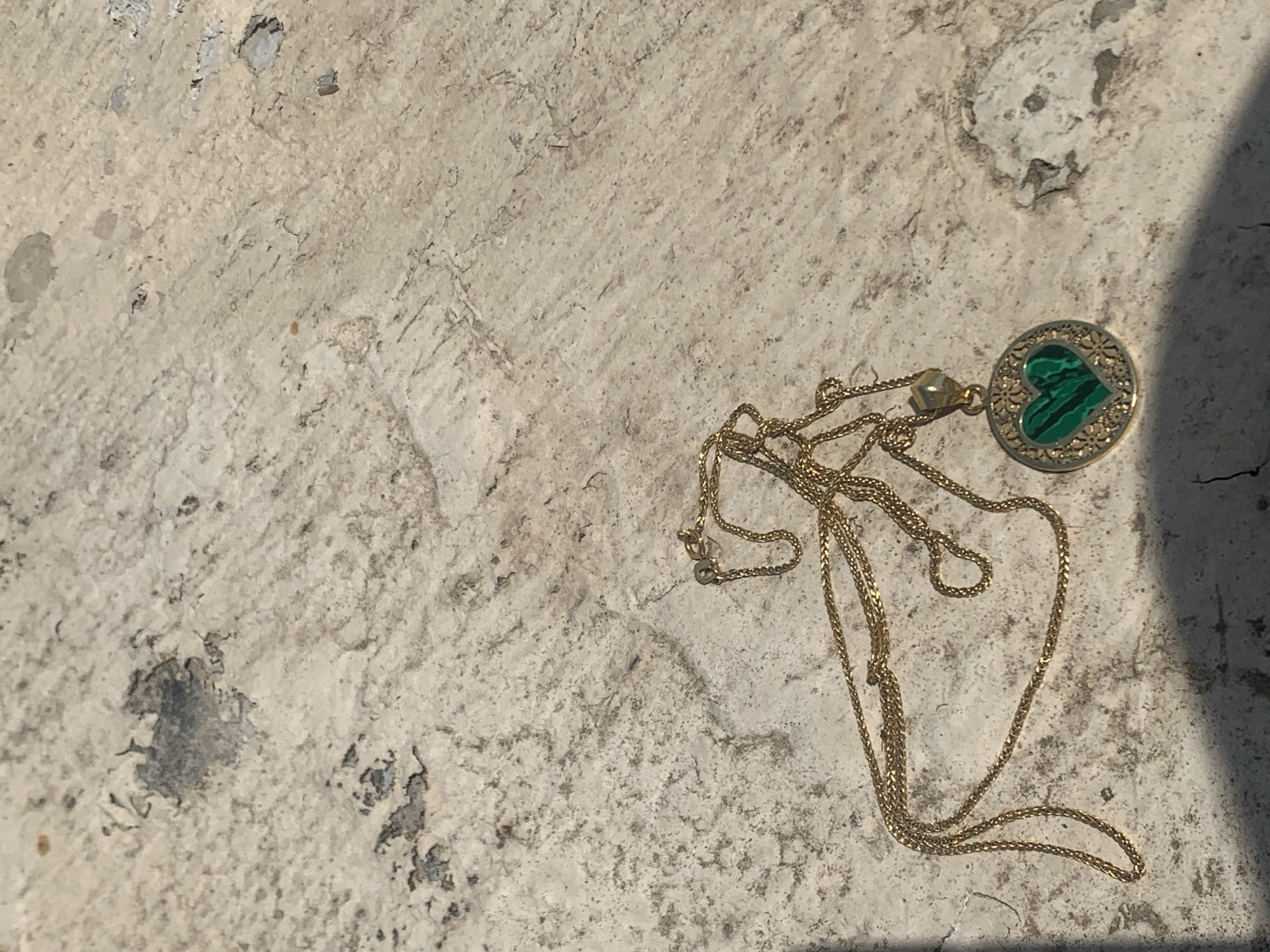 A close-up, high-angle shot shows a gold necklace with a circular pendant on a textured, light-colored surface. The pendant features an intricate gold design surrounding a green heart-shaped stone, possibly malachite. The necklace is draped across the surface, with the chain forming a loop. The surface appears to be stone or concrete, with rough textures and some darker, irregular patches. A shadow casts diagonally across the lower portion of the image, obscuring part of the surface and the necklace. There is no indication of people or activity in the image, and the scene is static. The location is identified as Khazar Raion, Azerbaijan.