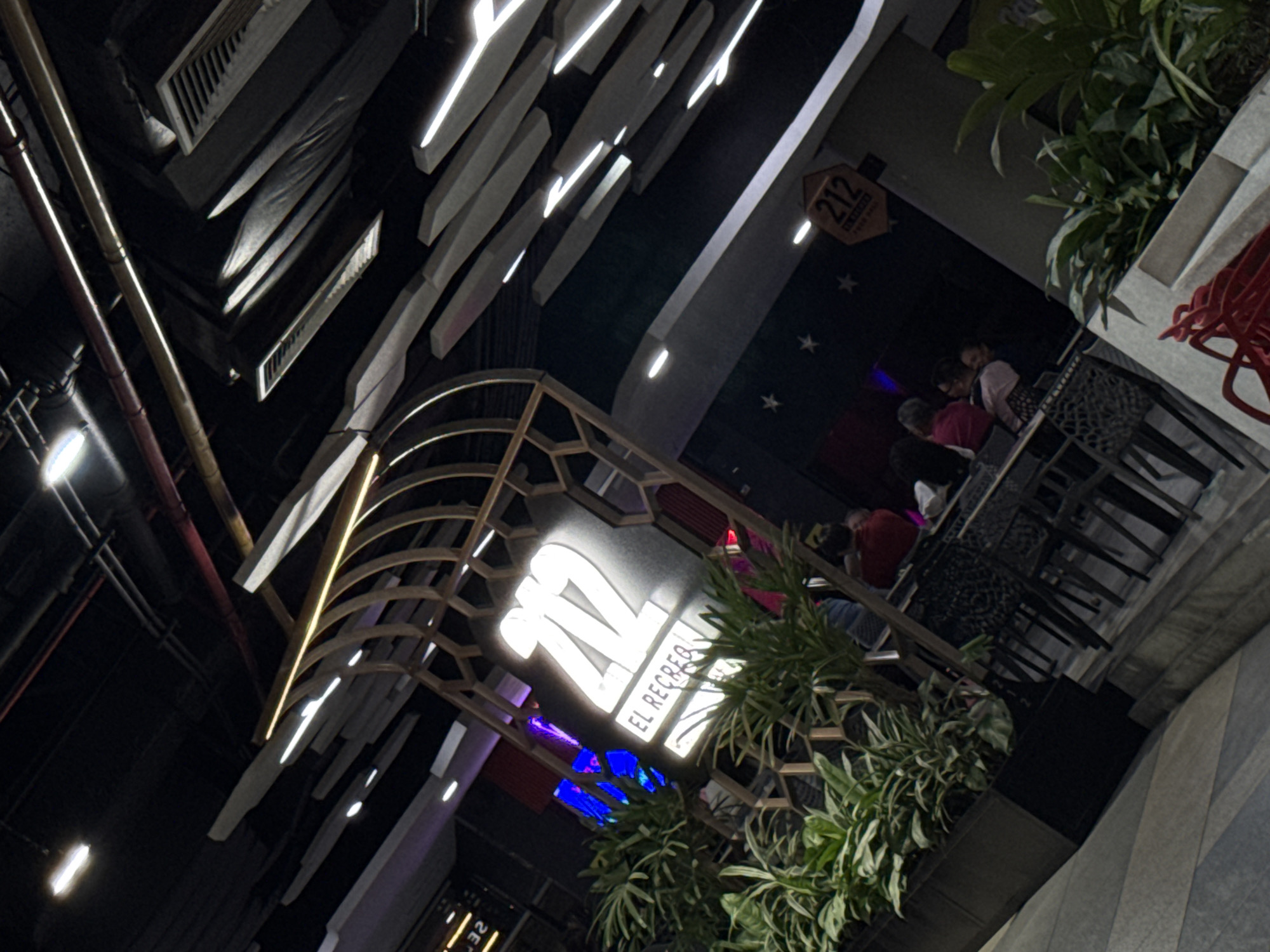 The image depicts a nighttime scene in Caracas, Venezuela, featuring the "212 El Recreo" restaurant. People are seated at tables on an outdoor patio. The building's exterior has a modern design with decorative lighting. The restaurant's sign is prominently displayed, and greenery surrounds the area.