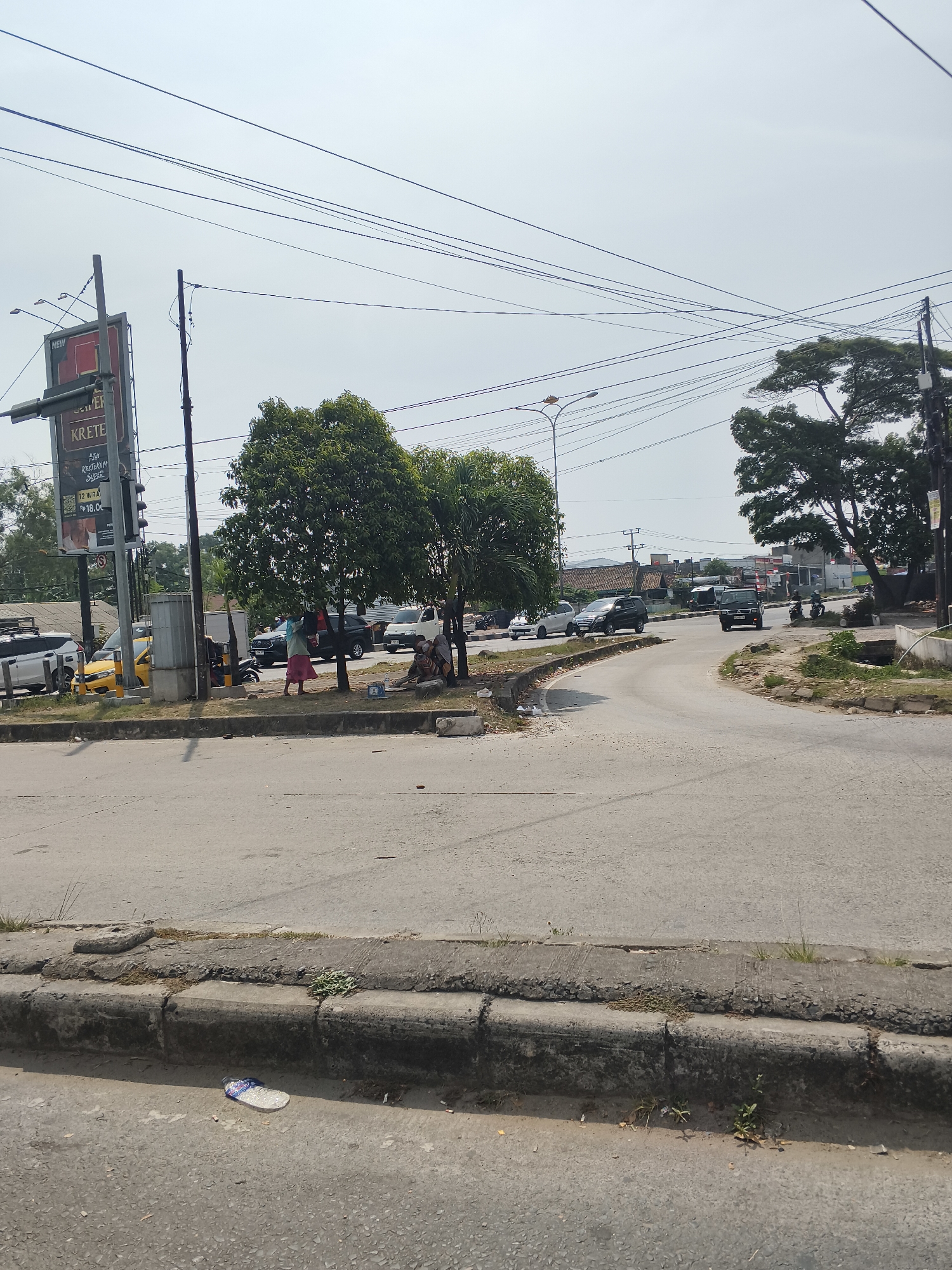This is a ground-level, eye-level, medium shot of a street intersection in Bandar Lampung, Indonesia, under a bright, overcast sky. The foreground shows a section of cracked concrete curb with some weeds growing through it, and a piece of white and blue plastic trash on the asphalt. A paved road curves away to the right, with a smaller paved area on the left. In the mid-ground, there is a yellow taxi, a white SUV, a black SUV, and a white van on the road. Several people are visible near the trees on the right side of the road. One person is walking away from the camera, and a group of people are sitting or squatting near a tree. A large billboard advertising "KRETEKNYA SUPER" is visible on the left side of the frame, with a traffic light above it. Overhead power lines crisscross the sky. Trees are present on both sides of the road, with one large tree on the right side partially obscuring the background. The background shows buildings and more trees, indicating a semi-urban environment.