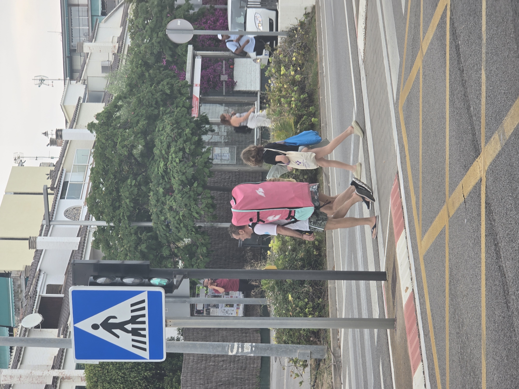 A pedestrian crossing on a sunny day in Sant Andreu de Llavaneres, Spain, shows two adults and a child walking across a road marked with yellow grid lines and a red and white curb. The older adult in the foreground is carrying a large, pink bag. A pedestrian crossing sign is visible, with a green traffic light illuminated. Buildings and vegetation line the background. A bus stop with seated individuals is visible in the midground.