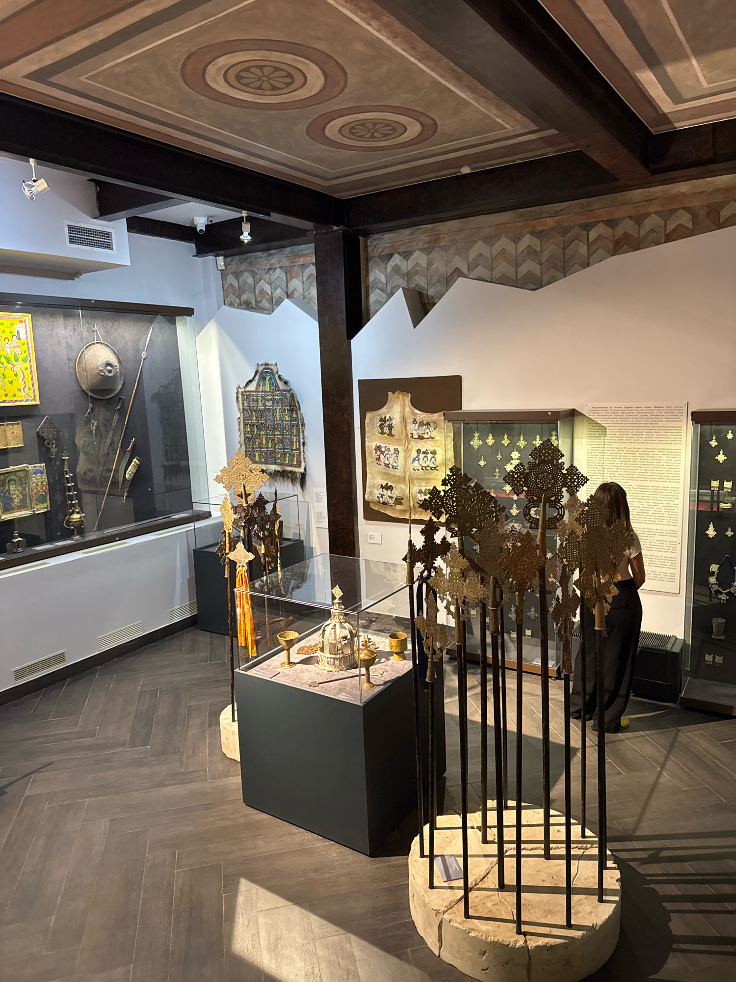 The image depicts an interior museum exhibit, likely focused on religious artifacts, possibly in Moscow, Russia. In the foreground, a collection of tall, ornate crosses made of a dark metal, possibly iron or bronze, are displayed on a rough-hewn stone base. A woman in dark pants and a white top stands near the crosses, looking at an informational display on the wall.
To the left, a glass display case contains various religious objects, including what appears to be a crown or ceremonial headdress, chalices, and other metalwork. Behind this, another glass case exhibits artifacts including a round shield, spears, and other weaponry. Further to the left, a framed religious painting is visible on the wall.
Behind the crosses, a wall features a tapestry-like textile with numerous small figures. Adjacent to this, a large informational plaque is mounted on the wall, with text in Cyrillic script. Other display cases with various golden crosses and jewelry are visible in the background.
The ceiling is ornately decorated with painted geometric patterns and circular motifs. The floor is tiled in a herringbone pattern with dark wooden planks. The lighting is a mix of ambient museum lighting and spotlights focused on the exhibits. The overall impression is of a well-curated historical or religious museum.
