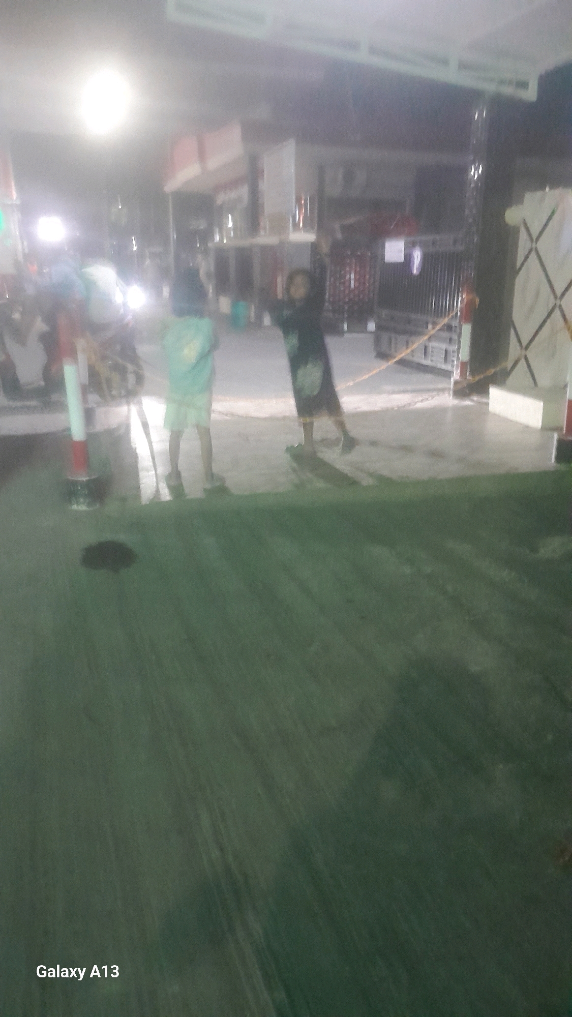 The image depicts a night scene on a paved surface, possibly a street or sidewalk, in Jekulo, Indonesia. Two young children are visible. The child on the left is wearing a light-colored, possibly green, shirt and shorts, and is facing away from the camera. The child on the right is wearing a dark-colored dress with a floral pattern and appears to be standing with arms raised, possibly dancing or playing. In the background, there are buildings with illuminated windows and structures that suggest commercial activity. A red and white striped pole is present to the left, and a chain barrier is visible in the middle ground. The lighting is dim, with some sources creating bright reflections on the wet or polished surface. The overall impression is of a casual, outdoor gathering or activity at night.