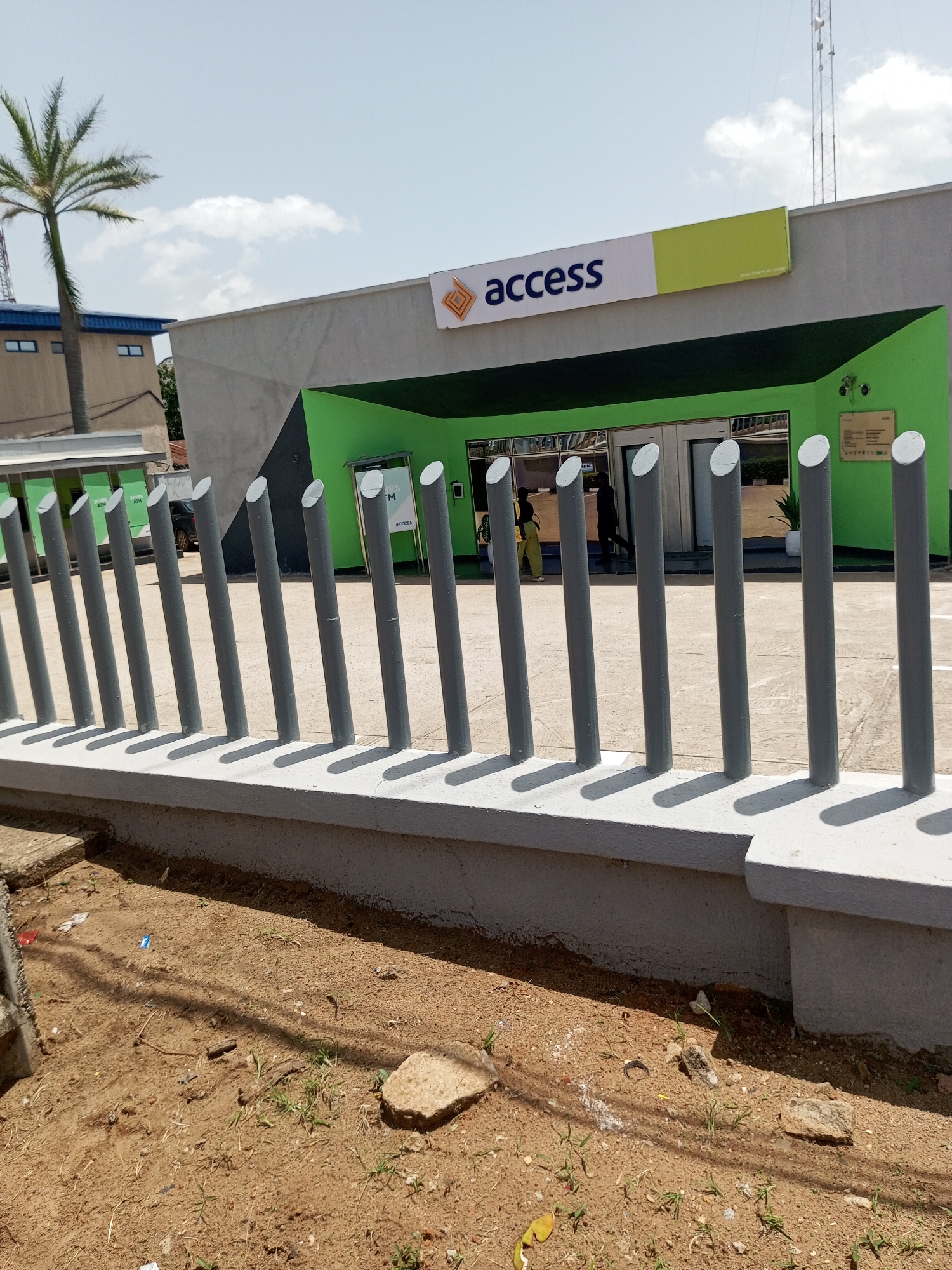The image depicts the exterior of an Access Bank branch in Bukuru, Nigeria, on a bright, sunny day. A row of grey metal bollards stands in the foreground, casting distinct shadows on the light-colored concrete ground. Behind the bollards, the bank building features a prominent green facade with a dark grey geometric pattern on the upper wall. The bank's signage, displaying "access" in dark blue letters alongside its logo, is visible above the entrance.
The entrance area of the bank is visible, with glass doors and windows reflecting the surroundings. Two people are seen near the entrance, one walking away from the camera and another partially visible inside the building. A potted plant adds a touch of greenery to the bank's exterior. To the left of the bank, a building with blue trim and windows can be seen, and a tall palm tree stands to the far left, its fronds reaching towards the sky. In the background, a tall radio or telecommunications tower is visible against the clear blue sky, which has a few wispy clouds. The ground in front of the bollards is sandy with scattered debris and small patches of dry grass. The overall impression is that of a typical urban commercial setting in Nigeria during daylight hours.