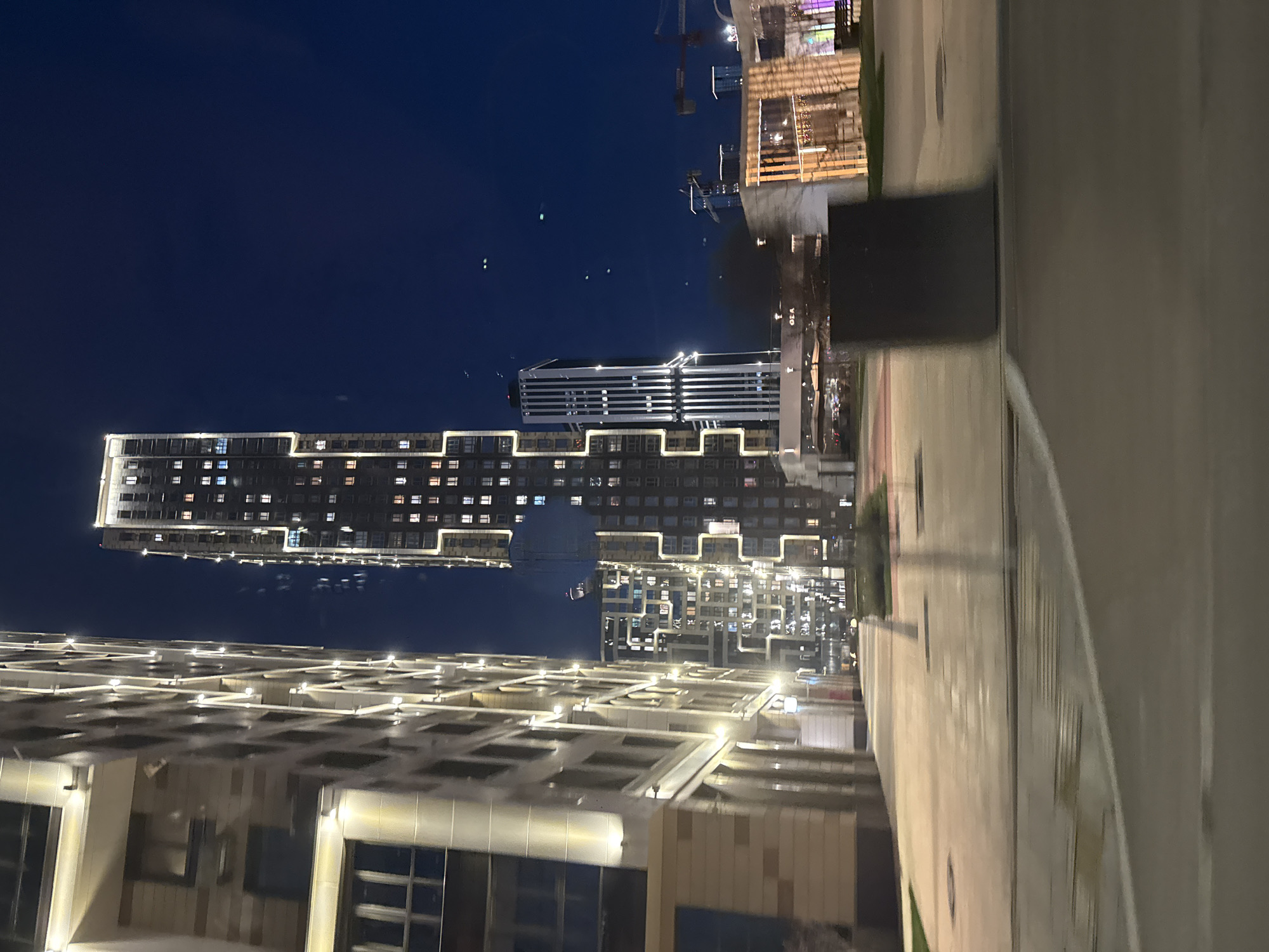 This media file captures a night scene in an urban environment, likely in Khimki, Russia, as provided by the context. The image is primarily composed of modern buildings illuminated against a dark blue night sky.
In the midground, a prominent, tall residential or office building stands, characterized by numerous square windows with many lights on, suggesting occupancy. The top perimeter of this building, as well as several horizontal lines along its facade, are outlined with bright, white strip lighting, creating a distinctive architectural feature. To the right and slightly behind this building, another tall structure with a more intricate, grid-like facade and similar lighting is visible. Further in the background, dimly lit construction cranes indicate ongoing development or industrial activity.
The foreground features a paved area, possibly a sidewalk or plaza, with a smooth, light-colored surface. To the left foreground, a lower-rise building with a textured, possibly ribbed, facade and external lighting is partially visible. The overall impression is one of a well-lit, contemporary urban landscape after dark. The sky is clear, with no visible clouds.
No people are visible in the image. The only discernable text appears in one of the cropped sections, showing "OBA" on a building or sign in the middle distance. The bright artificial lighting of the buildings strongly indicates it is nighttime.