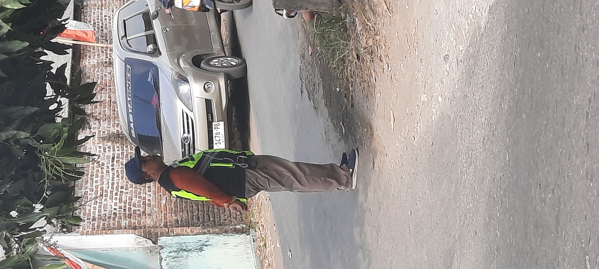 A man wearing a blue cap, an orange long-sleeved shirt, a black vest with neon green accents, and brown pants stands on a paved road in Jatitengah, Indonesia. His hands are clasped behind his back. He appears to be looking to his right. A silver car with a license plate reading "1476-PB" is parked on the right side of the road behind him. A motorcycle is partially visible on the far right of the image. In the background, a brick wall is visible, topped by a roof and green trees. An Indonesian flag is hanging on the right side of the building. The ground is covered in asphalt and some dry leaves.
