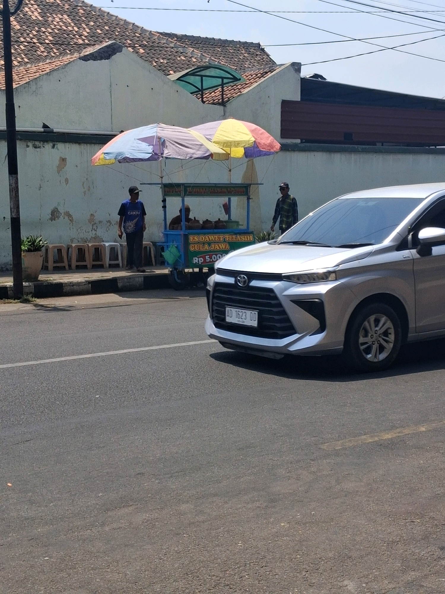 The image depicts a street scene in Surakarta, Indonesia. In the foreground, a silver Toyota Avanza car with license plate AD 1623 DD is parked on the right side of the road. The car is angled towards the viewer. Behind the car, a food cart with colorful umbrellas is set up on the sidewalk. The cart displays a sign that reads "ES DAWELL TELASIH GULA JAWA RP 5.000". Two individuals are visible near the cart. One man, wearing a dark blue t-shirt with the word "CAKRA" and dark pants, stands to the left of the cart. Another man, in a green and black checkered shirt and dark pants, stands to the right of the cart. Several small, light-colored stools are arranged on the sidewalk behind the man in the blue shirt. A potted plant is visible to the far left. The background consists of a light-colored building with a tiled roof. Overhead, electrical wires are strung across the sky. The scene appears to be illuminated by daylight.