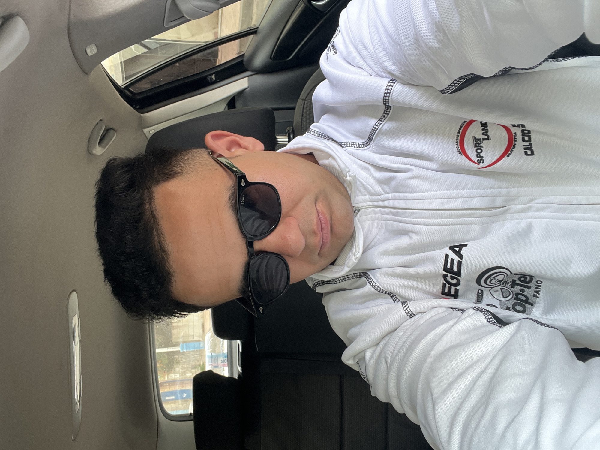 A middle-aged man with dark hair and sunglasses is sitting in a car with a sunroof. He is wearing a white zippered jacket with various sports logos and brands printed on it, including "SPORTLAND" and "EGEA." Outside the car window, there is a blurred cityscape with buildings and traffic. The photo was taken in Martil, Morocco.