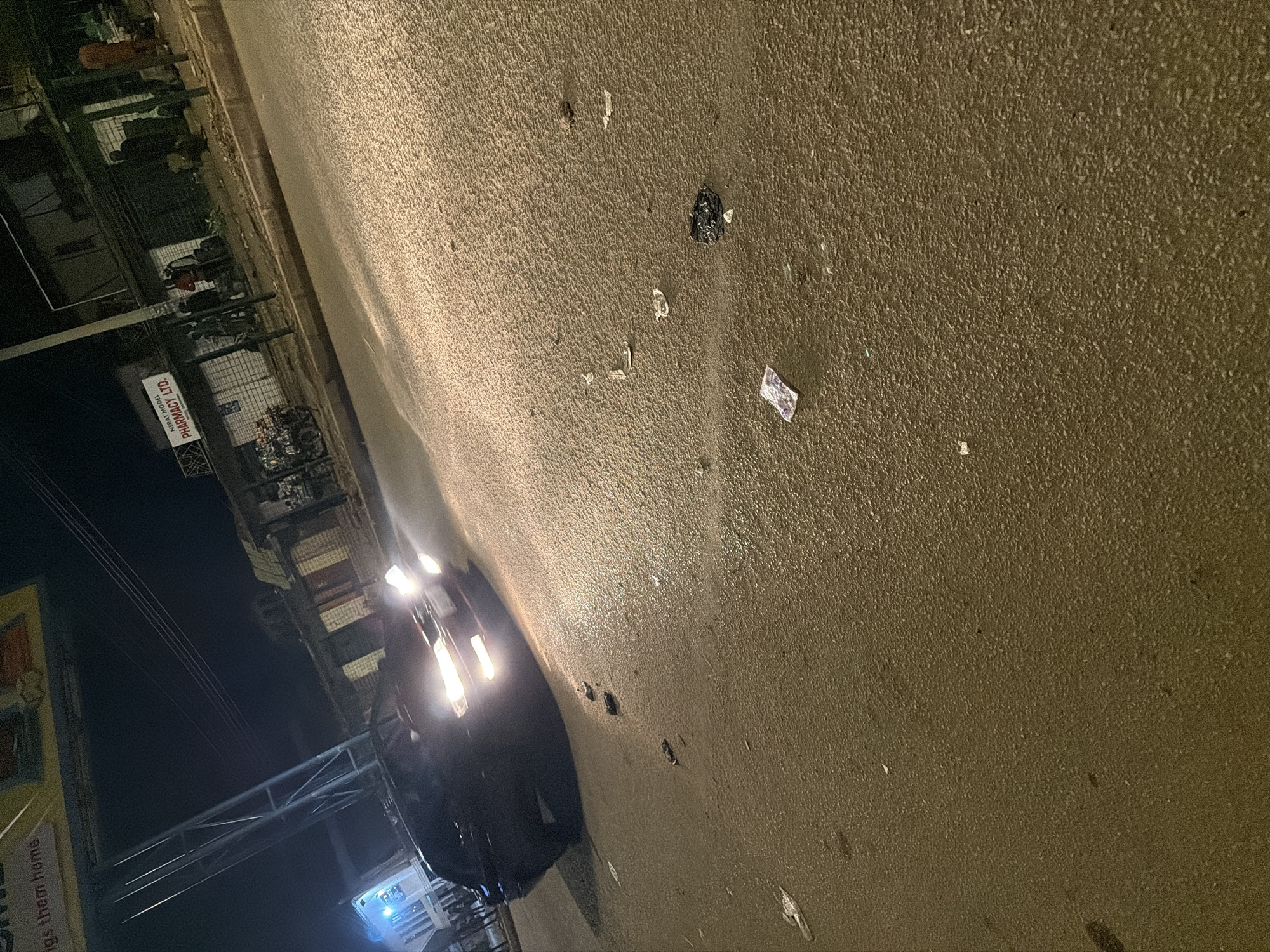In Bukuru, Nigeria, a nighttime street scene unfolds. A dark-colored sedan with its headlights on is positioned in the foreground, facing forward and slightly to the right. The car is on a paved road that appears wet, reflecting the light from its headlights and other ambient light sources. Scattered across the road are various pieces of debris, including what appears to be trash and possibly some paper currency.
In the background, a commercial building is visible, with a sign that reads "NERAY MODEL PHARMACY LTD." Several individuals are gathered outside the building, some standing, others appearing to be engaged in conversation. A bicycle with a large load on the back is parked on the side of the road. Power lines are visible overhead, and streetlights or building lights illuminate the scene, creating a contrast between illuminated areas and shadows. The overall impression is a busy street corner at night, possibly a market or commercial area.