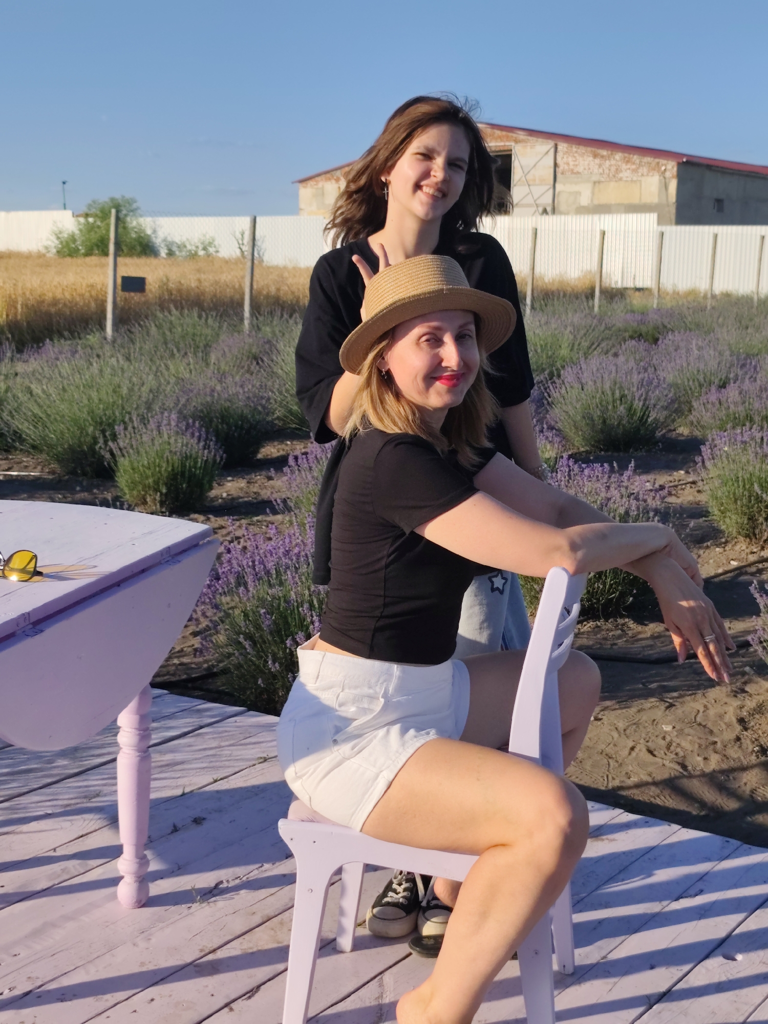 Two women are posing for a photo in a lavender field in Nechaiane, Ukraine. The woman in the foreground is sitting on a white chair, wearing a straw hat, a black t-shirt, and white shorts. She has bright red lipstick and is looking at the camera with a smile. Her arms are resting on the back of the chair. She is wearing rings on her left hand. The second woman is standing behind her, wearing a black shirt and smiling at the camera. She has a cross necklace and is making a peace sign with her right hand.
To the left of the women is a small, light purple table with yellow sunglasses on it. The table and chair are on a light pink wooden deck. Behind the deck is a field of lavender. In the background, there is a building with a red roof and a white fence. The sky is clear and blue, and the sun appears to be setting, casting long shadows. The overall mood is cheerful and relaxed.