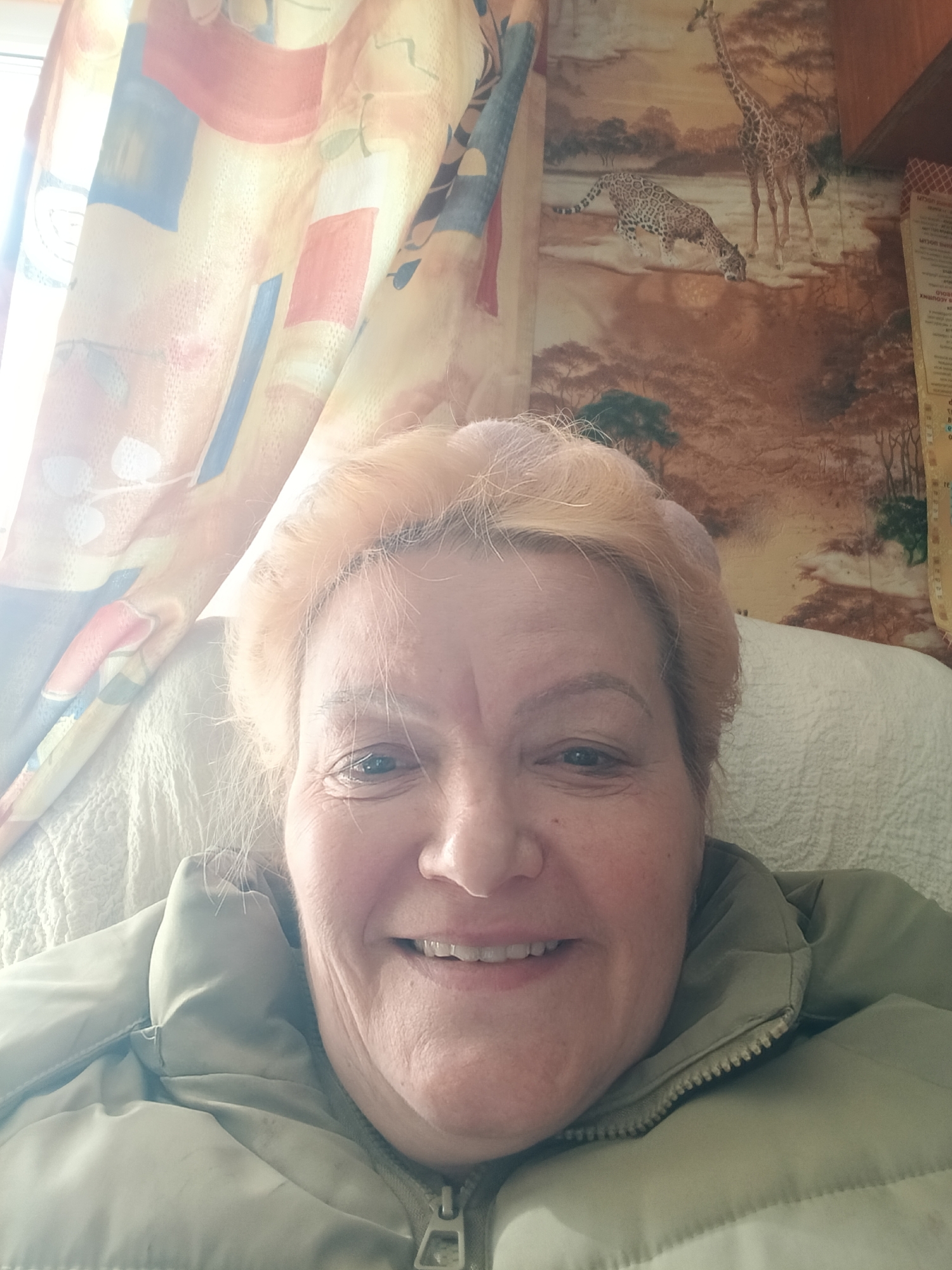 A smiling middle-aged woman with light blonde hair is captured in a close-up selfie, looking directly at the camera. She appears happy and relaxed. She is wearing a puffy, olive-green zippered jacket. Bright sunlight streams in from the left, illuminating translucent curtains behind her that feature a colorful, abstract pattern with red, blue, and yellow geometric shapes.
To the woman's right, the background reveals a section of wallpaper depicting an African safari scene. A spotted big cat, possibly a leopard or jaguar, is shown bending down to drink from water, while a tall giraffe stands in the background. Sparse trees characteristic of a savannah landscape are also visible. A light-colored, quilted pillow or cushion supports her head.
On the far right edge of the image, a piece of paper with visible Cyrillic text is partially obscured, suggesting it might be a calendar or schedule. Readable fragments include days of the week: "ВТОР" (Tuesday), "СРЕДА" (Wednesday), "ЧЕТВЕРГ" (Thursday), "ПЯТНИЦА" (Friday), "СУББОТА" (Saturday), and "ВОСКР" (Sunday). The bright light indicates it is daytime. The setting is an indoor space, likely a home, in Красный Маяк, Russia.