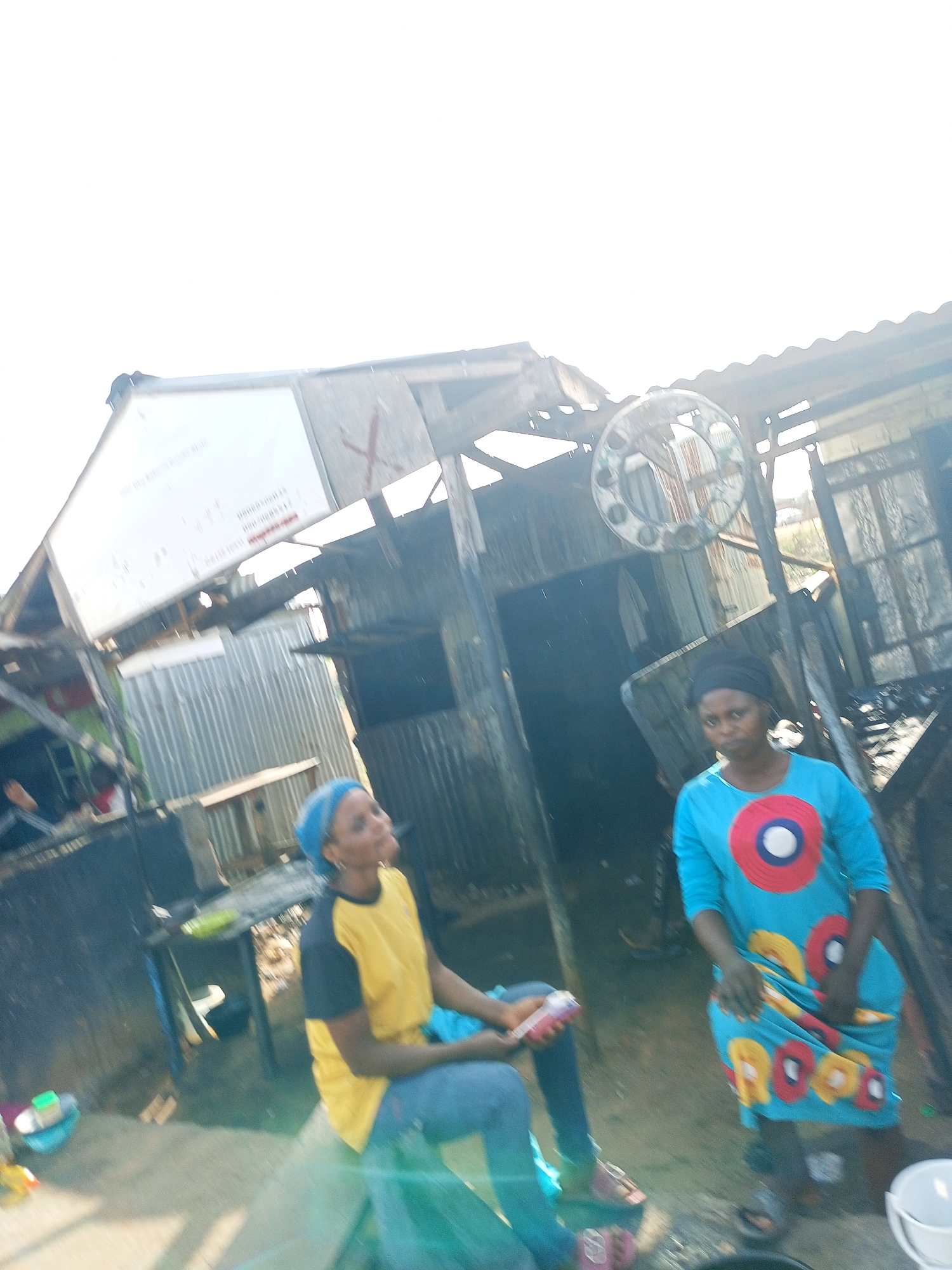 In Gombe, Nigeria, two women are pictured outside a makeshift structure. The structure appears damaged, with corrugated metal walls and a partially collapsed roof. A large white sign with red lettering is affixed to the upper portion of the structure. The woman on the left, wearing a blue headscarf and a yellow and black t-shirt, sits on a bench or ledge, holding a pink object. The woman on the right, wearing a black headscarf and a blue dress adorned with colorful circular patterns, stands facing forward. The ground is dirt and debris. The overall scene suggests an outdoor market or informal settlement.