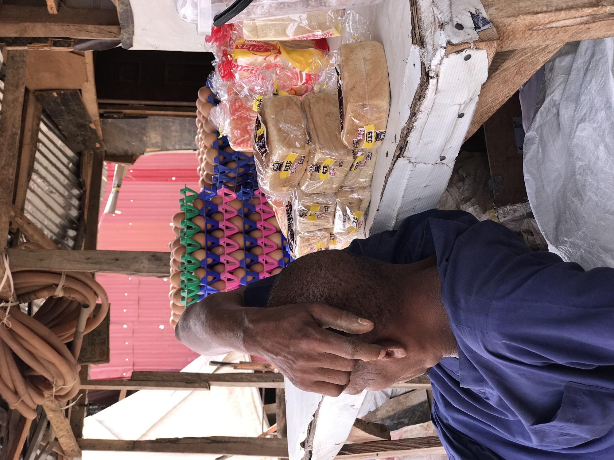 This vertical shot captures a bustling market scene, likely in Abuja, Nigeria, focusing on a stall selling essential goods.
In the foreground, a person with dark skin and short hair is visible from the side, wearing a dark blue long-sleeved shirt. Their right hand is raised to their face, covering the eye area, possibly shielding from bright light, or engaged in a private moment of reflection or weariness.
To the left of the person, a market counter displays a variety of goods. Numerous brown eggs are neatly stacked in colorful plastic trays (green, blue, and pink), indicating a substantial stock. Adjacent to the eggs, several loaves of packaged bread are arranged. Each loaf is sealed in clear plastic, and many feature distinct yellow labels with the visible text "GTC Bakery Bread," identifying the product and brand.
The background and surrounding environment suggest a rustic, outdoor market setting. Wooden beams and supports form the structure of the stall. A section of pink corrugated metal roofing is visible, providing shelter from the elements. On the far left, coils of brown hoses or cables are suspended, tied with white string, hinting at various utilities or items for sale. On the right, a large, light-colored tarpaulin or cloth hangs, likely offering additional shade.
The overall impression is a vibrant, everyday market in an African urban setting, captured during daylight hours. The specific details, such as the packaged bread with local branding, reinforce the location context of Abuja, Nigeria.