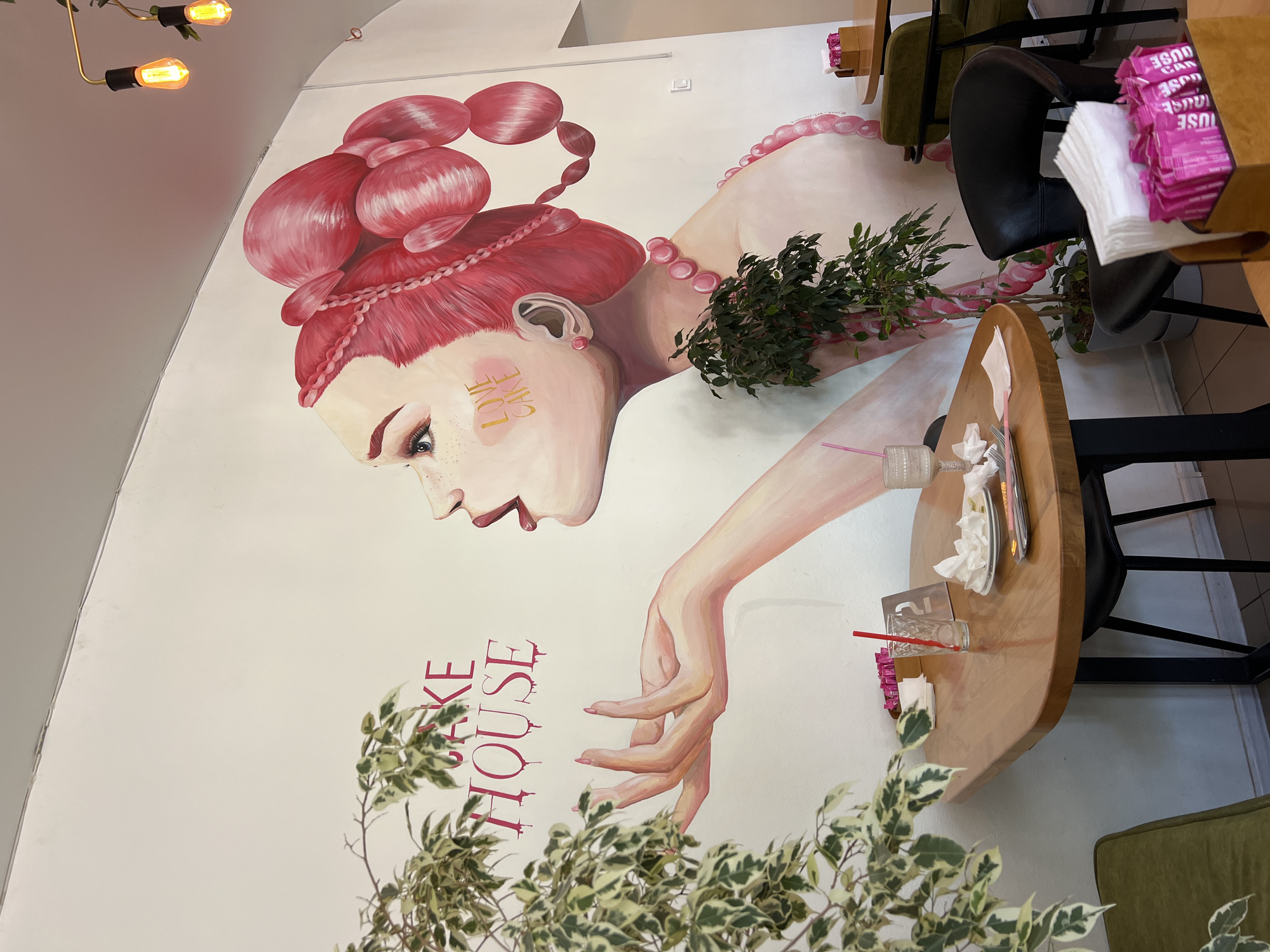 The image depicts the interior of a cafe in Odesa, Ukraine, with a large mural covering the white wall. The mural features a stylized portrait of a woman with pink hair, blowing a kiss, and the words "CAKE HOUSE" are painted in pink above her. The woman is adorned with pink pearls. In the foreground, a wooden table is set with a glass of water, napkins, and cutlery, suggesting a recent meal. A green upholstered chair is visible to the right. A potted plant with variegated leaves stands in front of the mural. Various lighting fixtures are visible on the ceiling.