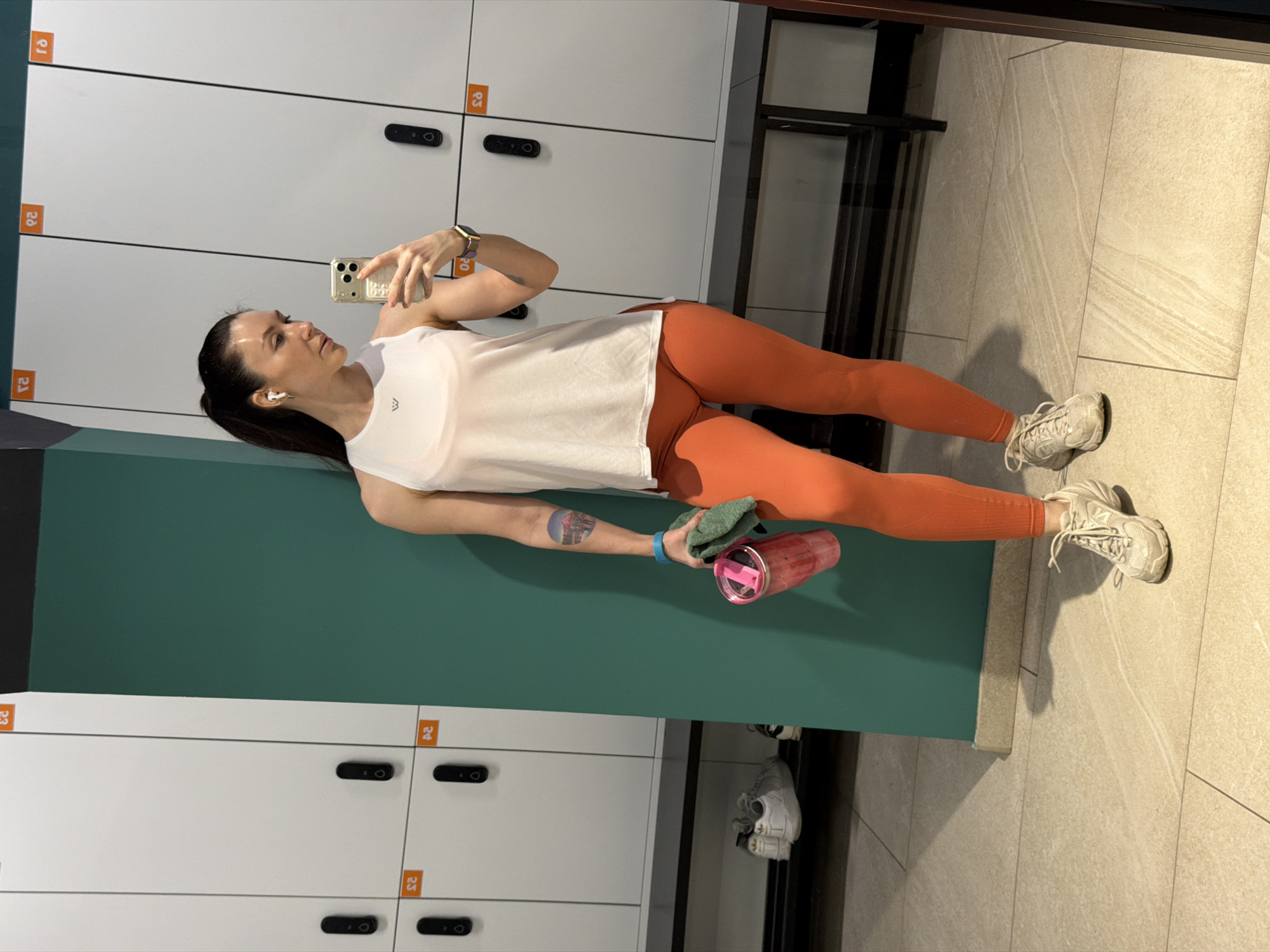 A full-body mirror selfie captures a woman in what appears to be a gym changing room or locker area in Perm, Russia. The woman, with dark hair tied back, stands facing left, holding a light-colored smartphone in her right hand to take the picture. She wears white wireless earbuds, a gold-toned watch on her left wrist, a white sleeveless athletic top, bright orange form-fitting leggings, and white sneakers. A small circular tattoo is visible on her left forearm. In her left hand, she holds a dark green towel and a pink and clear water bottle.
The setting consists of white metal lockers with black handles/locks lining the wall behind and to the left of her. To her right and in front, there is a teal-colored partition or wall. The floor is covered in light-toned rectangular tiles. The overall environment appears clean and well-lit by artificial lighting.
Notable details include orange square stickers on the lockers displaying numbers that resemble '23', '24', '28', and '29', rendered in a style similar to Devanagari script. A small, dark, stylized "M" logo is visible on the front of her white top. The woman's expression is neutral and focused. The image provides no specific cues for the time of day or outdoor weather.