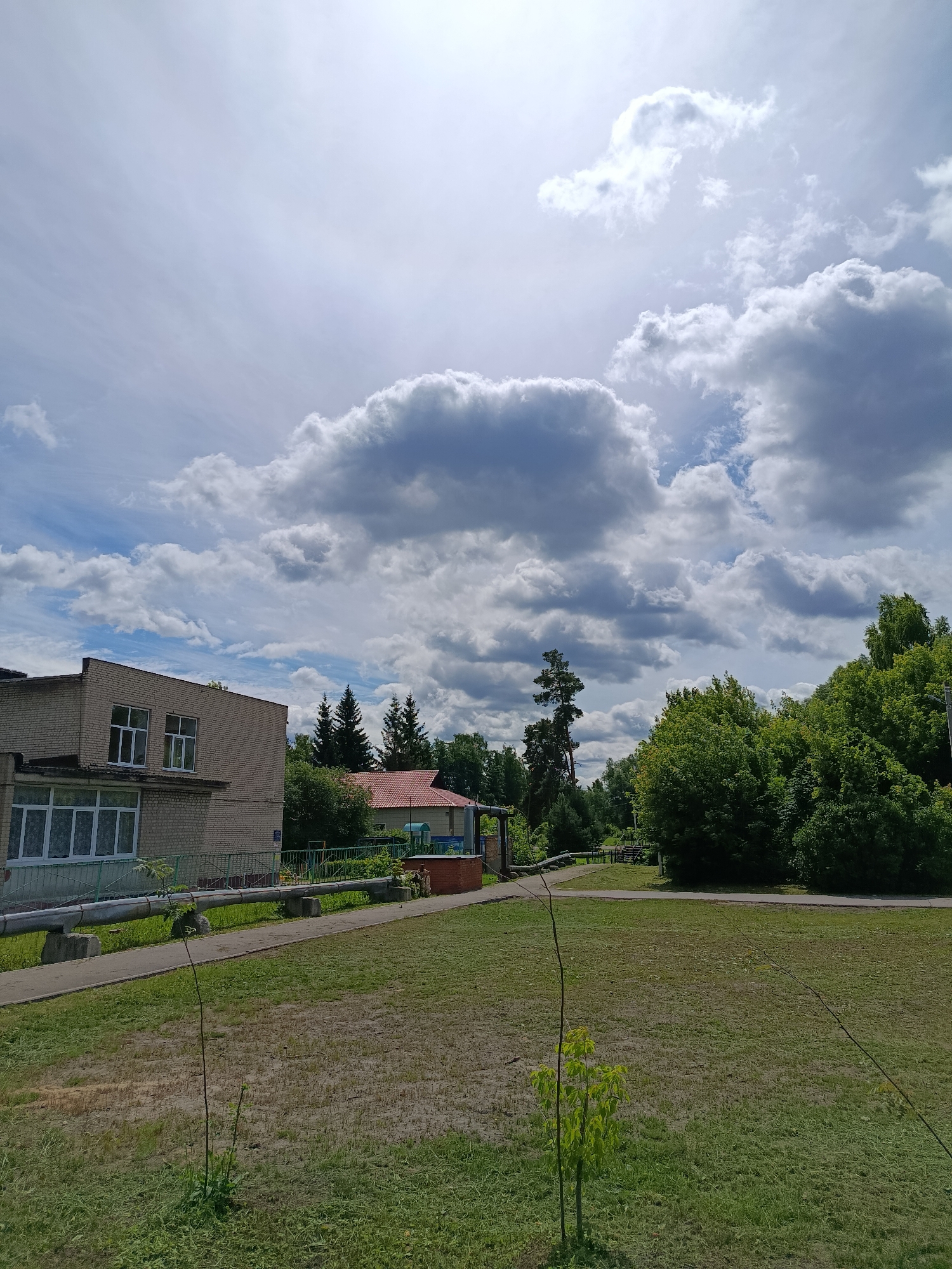 The image captures a daytime scene in Vereya, Russia, featuring a low-rise brick building on the left, with multiple windows, some of which are large and boarded up with white frames. In front of the building, a paved pathway runs parallel to a large, silver pipe supported by concrete blocks. The ground slopes away from the pathway and is covered in mown grass and patches of bare earth. Beyond the building and pathway, a red-roofed structure is visible, with fencing and mature trees in the background. The sky is dominated by dramatic, heavy clouds, interspersed with patches of blue sky, and the sun is partially obscured. The overall impression is one of a slightly overcast but bright day in a semi-urban or suburban setting with abundant greenery.