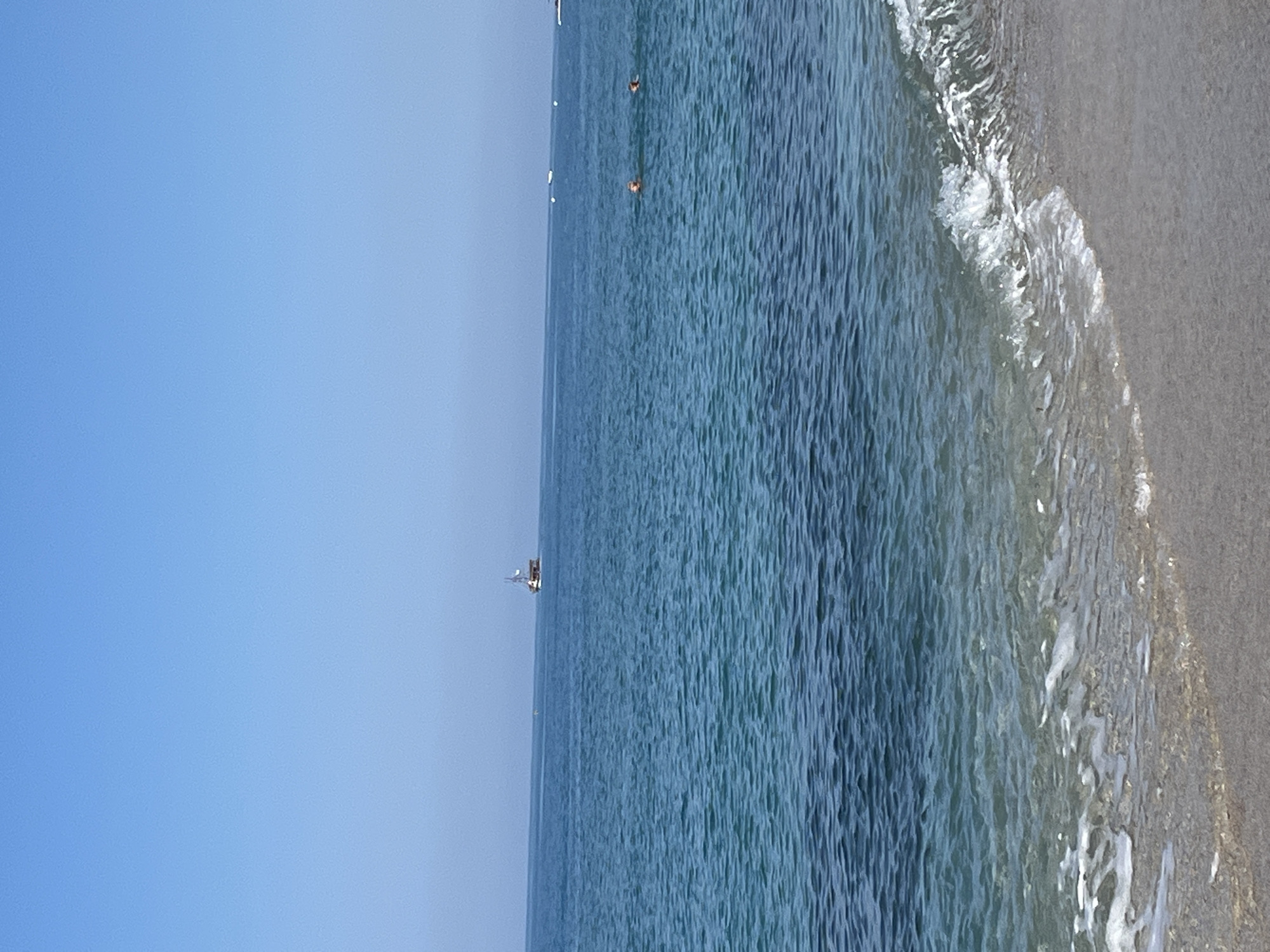 The image depicts a beach scene in Alanya, Turkey. A light blue sky fills the left portion of the frame. The sea occupies the central and right sections, transitioning from deep blue in the distance to lighter shades near the shore. There are a few small, indistinguishable figures visible in the water, and a small boat is visible on the horizon. The shoreline is a light gray color and shows the wash of waves. The image appears to be rotated, as the horizon is vertical rather than horizontal.