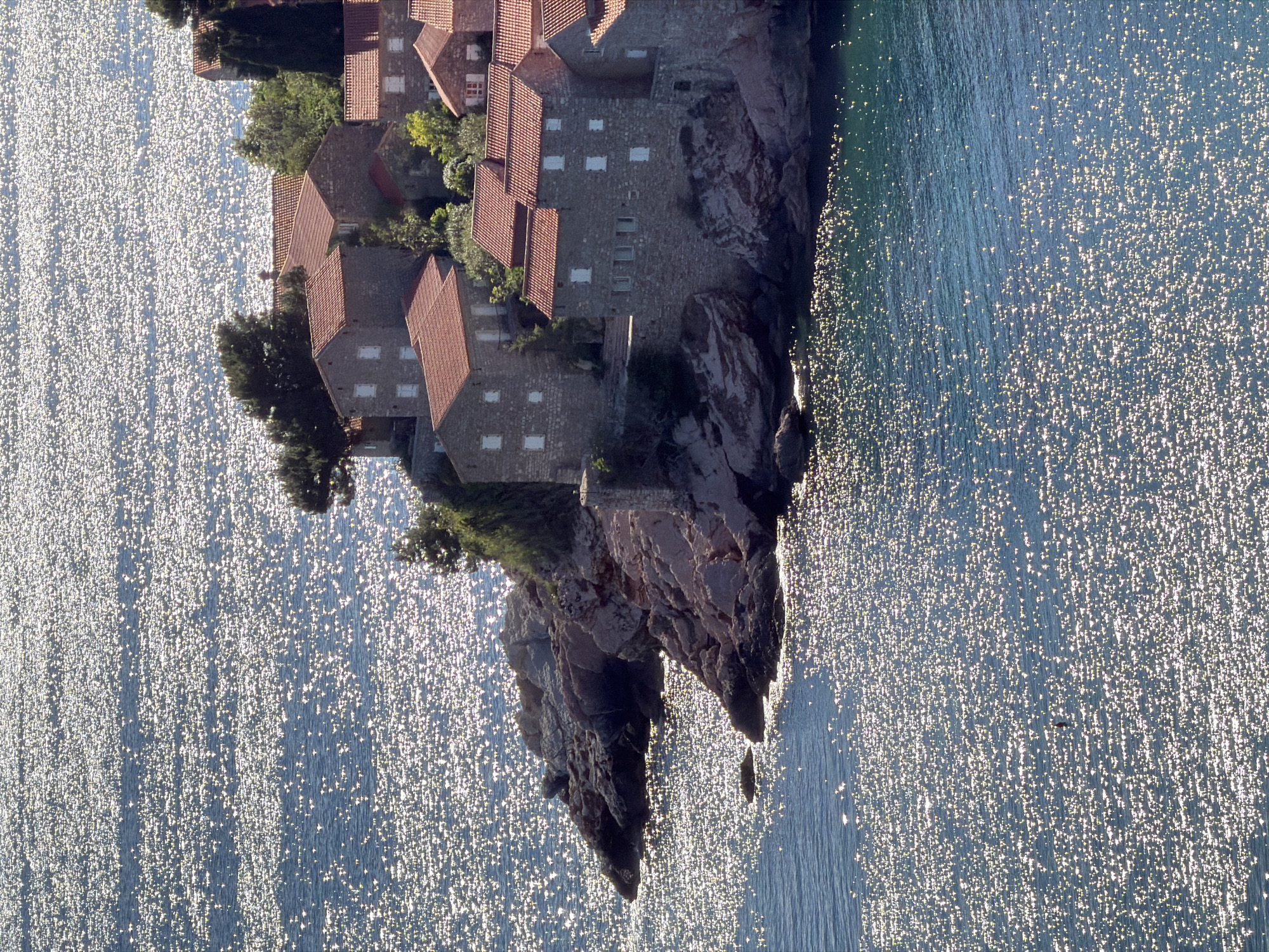 This image depicts Sveti Stefan, Montenegro, an island resort village characterized by terracotta-roofed buildings clustered on a rocky peninsula. The camera angle appears to be from an elevated perspective, showcasing the village in contrast with the surrounding Adriatic Sea. The sea's surface is illuminated by sunlight, creating a sparkling effect. The rocky coastline on which the village is built is visible, with a mix of natural rock formations and man-made structures that constitute the resort. There are no people visibly identifiable in the image.
