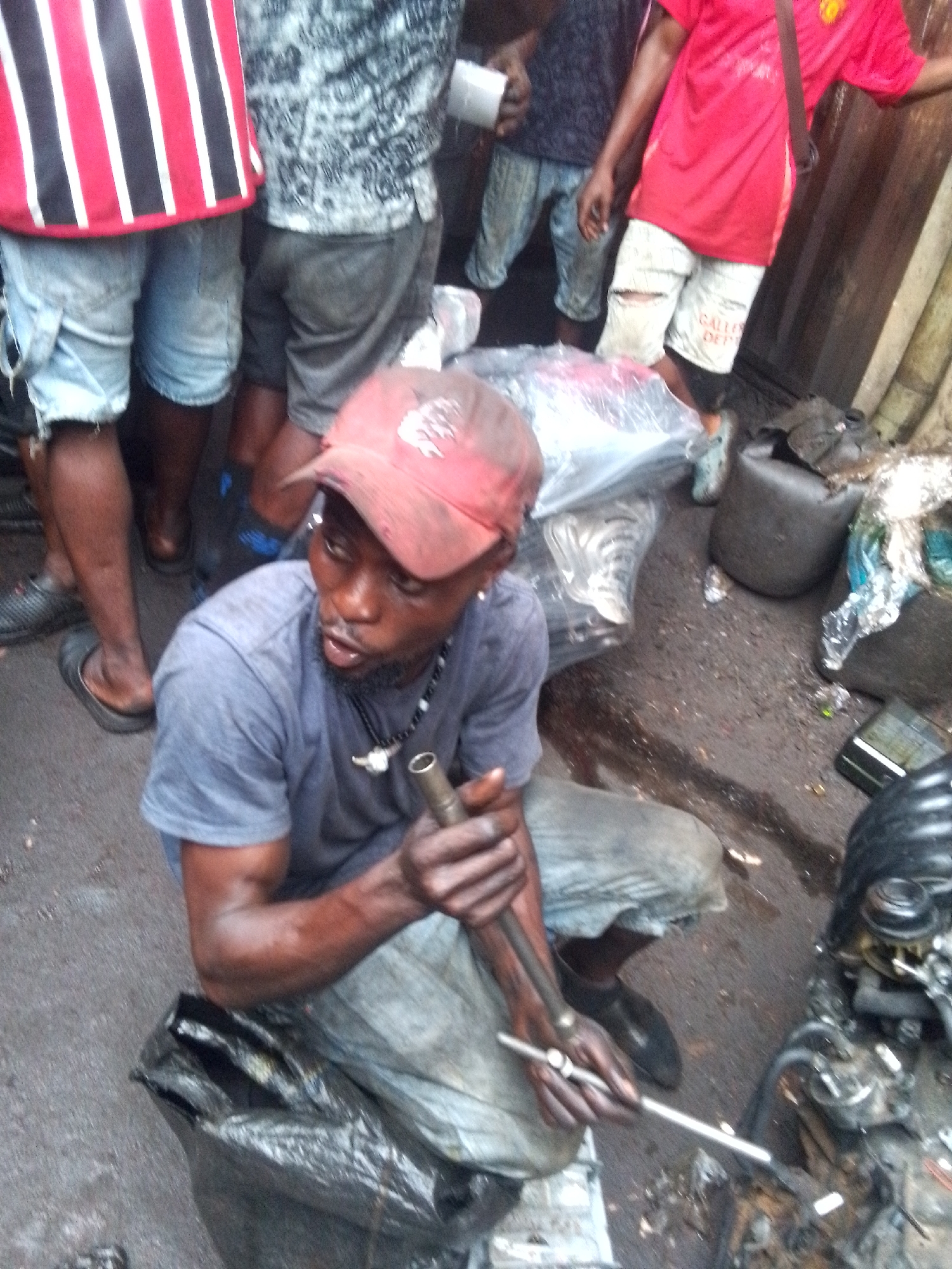 The photo depicts a mechanic working on a car engine in Onitsha, Nigeria. The mechanic is a dark-skinned man wearing a red baseball cap, grey t-shirt, light grey trousers, and a necklace. He's kneeling on a black plastic sheet while holding a long metal bar in his hands, presumably a tool to work on the engine. The engine is on the ground next to him. Other people are standing around in the background. The ground is wet, and there are containers scattered around. The environment appears to be an open-air space, possibly a roadside or a market area.