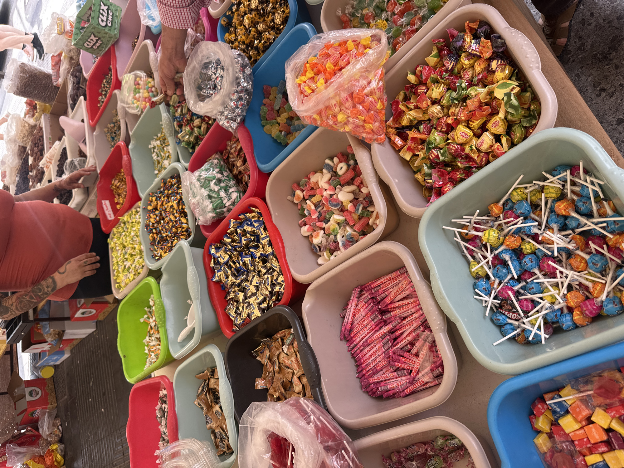An overhead, vibrant shot captures a market stall overflowing with an extensive array of bulk candies displayed in numerous plastic containers of various colors, including red, green, blue, and beige. The scene is bustling with commercial activity, featuring at least two individuals, indicated by visible hands. One person's arm, adorned with tattoos, reaches into a bin of yellow and white wrapped candies, while another hand is visible near a bag of silver-wrapped sweets.
The stall offers a wide variety of confectionery, including colorful lollipops in shades of blue, pink, yellow, and orange, many with white sticks, some with "KOP" visible on their wrappers. There are bins overflowing with individually wrapped hard candies, gummy sweets of different shapes and colors (some sugar-coated), and various types of chocolates and other sweets in shiny, metallic wrappers. A large clear plastic bag holds a mound of orange and yellow hard candies.
Among the visible text on the packaging, sticks of pink-wrapped sweets bearing the text "PALOTES" are prominently displayed in one bin. Other wrappers show the brand names "GERIO," "CLIX ONE" on a green box, and "Fini" on smaller wrappers. The abundance and colorful display suggest a lively, inviting atmosphere for purchasing sweets, likely in a market or specialty candy store. The lighting appears consistent, suggesting an indoor or covered outdoor setting, with no specific time of day discernible.