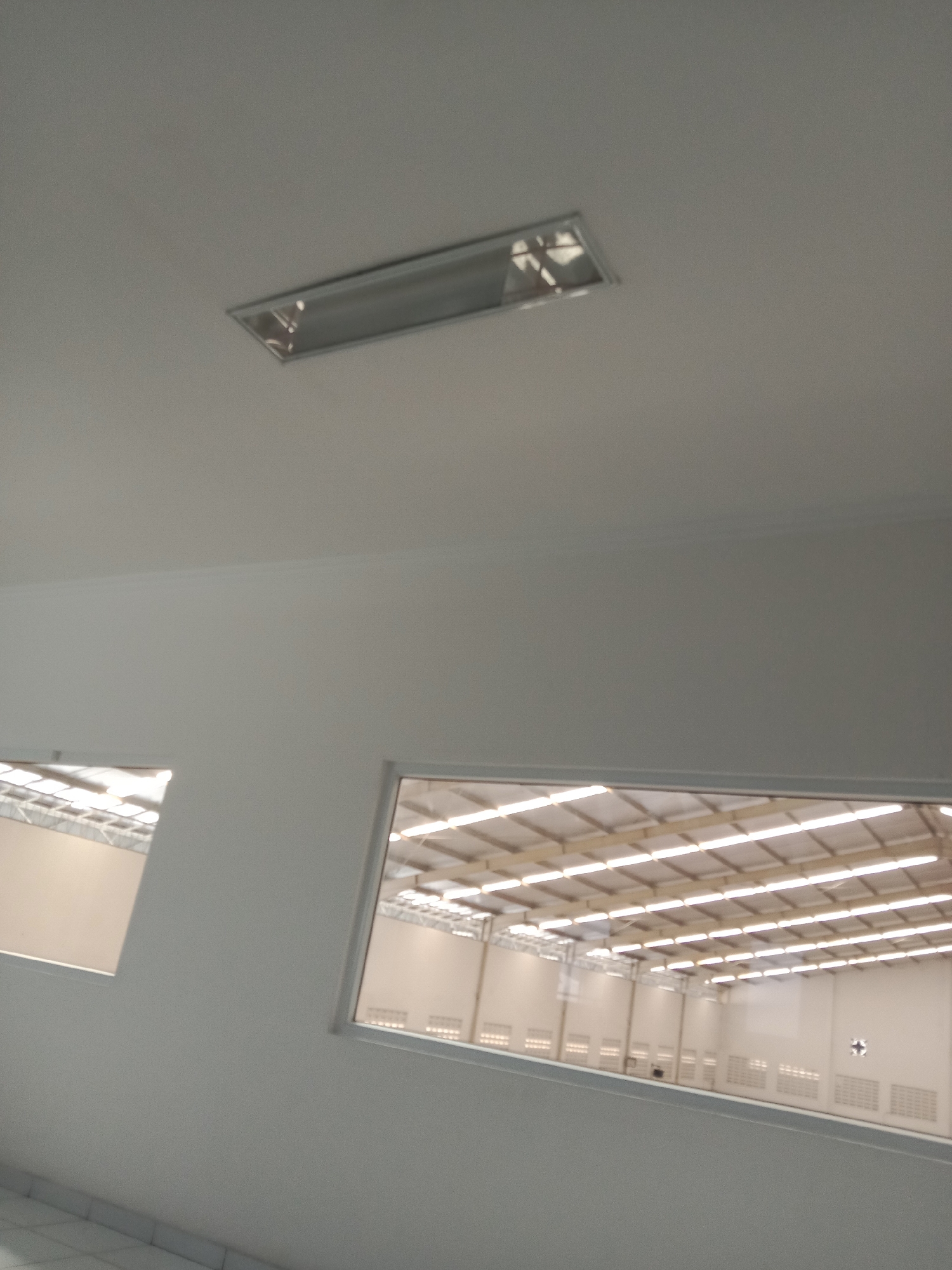 The image depicts the interior of a modern, empty warehouse or industrial building. A rectangular fluorescent light fixture is mounted on the white ceiling. Two large, horizontal windows or openings in the white wall offer a view into another section of the building. Through these openings, rows of bright, parallel lights illuminate the corrugated metal ceiling structure of a larger space. This space appears to be a vast, empty industrial hall with white walls. The floor visible in the foreground is tiled. The scene is clean and well-lit, suggesting a functional and maintained facility. The visual style and architecture are consistent with contemporary industrial designs commonly found in areas like the industrial estates on the outskirts of Semarang, Indonesia, known for their manufacturing and logistics operations.