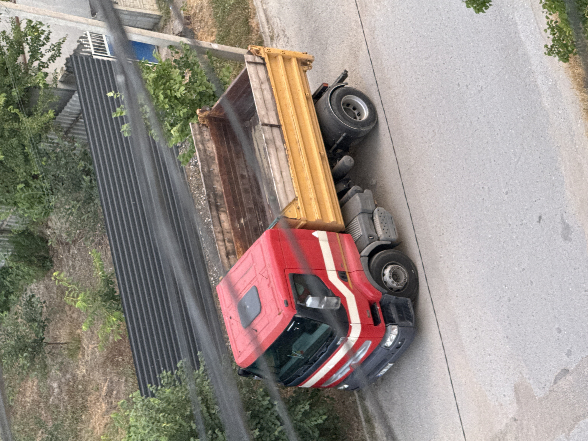 A red and yellow dump truck is parked on the side of a paved road in what appears to be a rural or suburban setting. The truck has a yellow, empty dump bed and a red cab with white stripes. The image is taken from an elevated viewpoint, looking down at an angle. Trees and some buildings, including a dark, corrugated metal structure, are visible in the background. The road surface is rough and speckled. The lighting suggests it is daytime, possibly late afternoon, with the sun casting some shadows. There are power lines or cables running across the foreground, slightly obscuring the view of the truck and the surroundings. No people are visible. The overall impression is of a parked construction vehicle in a somewhat unkempt environment.