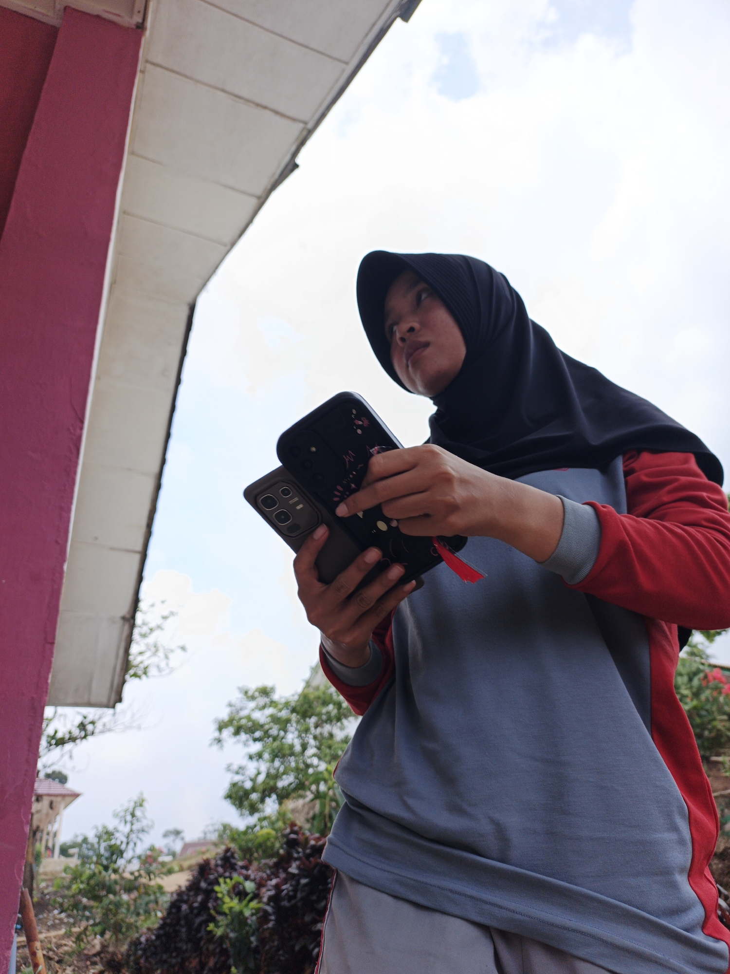 A low-angle medium shot captures a person wearing a black hijab and a red and gray long-sleeved shirt, holding two smartphones in their hands. The person is outdoors, with a pink wall and a white overhang of a building on the left. In the background, a cloudy sky is visible, along with trees and bushes. The person's gaze is directed upwards and to the right, with their lips slightly parted. One of the phones appears to be a flip phone, while the other is a dark-colored smartphone with a floral pattern on its case. The setting suggests a residential or semi-public area, possibly in Jepara, Indonesia, given the greenery and architectural hints. The lighting is bright and overcast, indicating daytime.