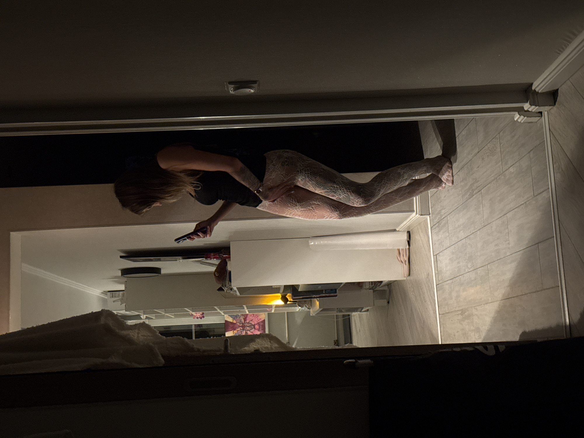 A woman is standing in a dimly lit room, illuminated by a phone light she is holding. She is wearing a black tank top and white patterned tights. Her left hand is resting on her thigh, and she appears to be looking at her phone. In the background, there is a white shelf unit with various items on it, including books and a lamp. There is also a framed piece of art on the wall. The room has tiled flooring. The scene is indoors, and there is no clear indication of the location within Moscow, Russia.
