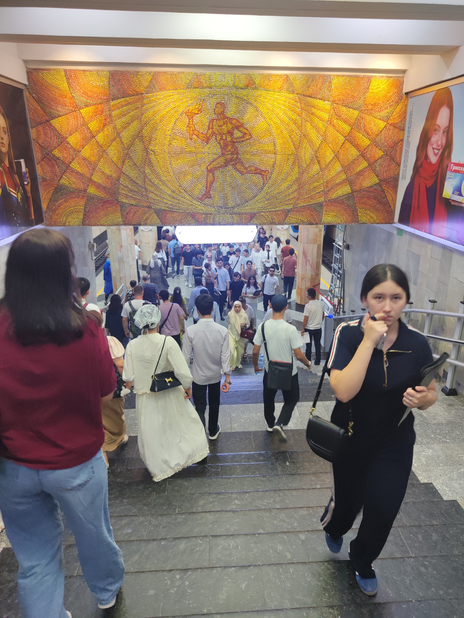 The image shows an underground subway station in Tashkent, Uzbekistan, with people descending stairs. A large mosaic adorns the wall above the stairs, depicting a running figure with a torch. Posters of women are visible on either side of the mosaic. The people are dressed in casual clothing, with some women wearing hijabs. The station appears to be busy, with many individuals walking down the stairs. The architecture suggests a Soviet-era design, common in many former Soviet republics. The lighting is bright, illuminating the mosaic and the crowd. The overall scene is one of daily transit in a public space.
