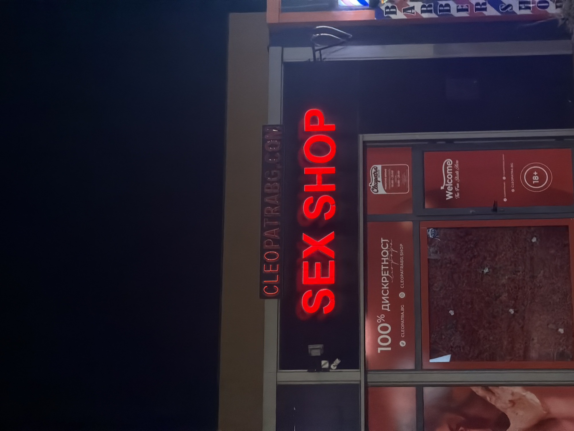 The image shows the exterior of a store with neon signage. A large red neon sign in the center of the image reads "SEX SHOP". Above this, a smaller red neon sign reads "CLEOPATRA BG.COM". To the left of the "SEX SHOP" sign, there is a banner with the text "100% дискрETHOCT" and two web addresses, "CLEOPATRA.BG" and "CLEOPATRABG.SHOP". To the right of the "SEX SHOP" sign is a door with a sign that says "Welcome The Fun Starts Here" and an "18+" icon. The building appears to be at street level, and the signage suggests it is a retail establishment. The overall impression is that of a business operating in an urban environment at night.