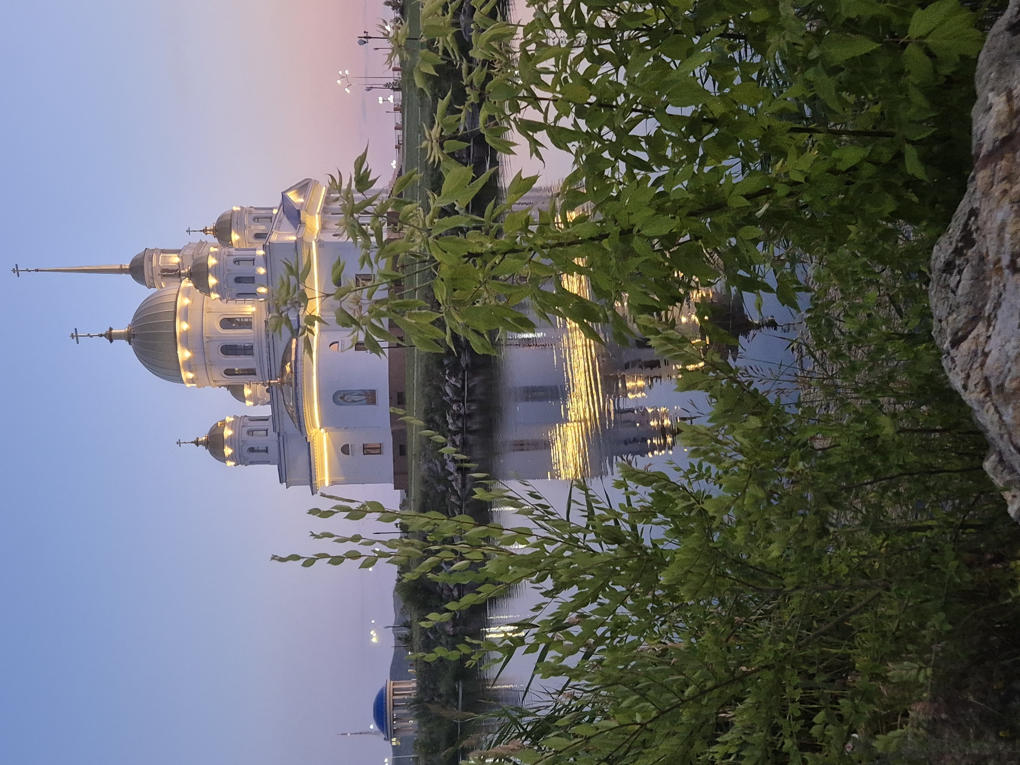 The image depicts the Assumption Cathedral, a prominent landmark in Kokshetau, Kazakhstan, at dusk. The white cathedral, adorned with gold domes and illuminated by warm lights, stands majestically on a hill overlooking a tranquil body of water. The water reflects the cathedral's golden glow and the twilight sky, creating a serene and picturesque scene. Lush green foliage in the foreground partially obscures the view, adding a naturalistic element to the composition. In the distance, a smaller blue-domed building and streetlights suggest the presence of the surrounding city. The sky transitions from a pale blue to soft hues of pink and orange, indicating the end of daylight. The scene is devoid of any people or animals, emphasizing the architectural beauty and the peaceful ambiance of the location.