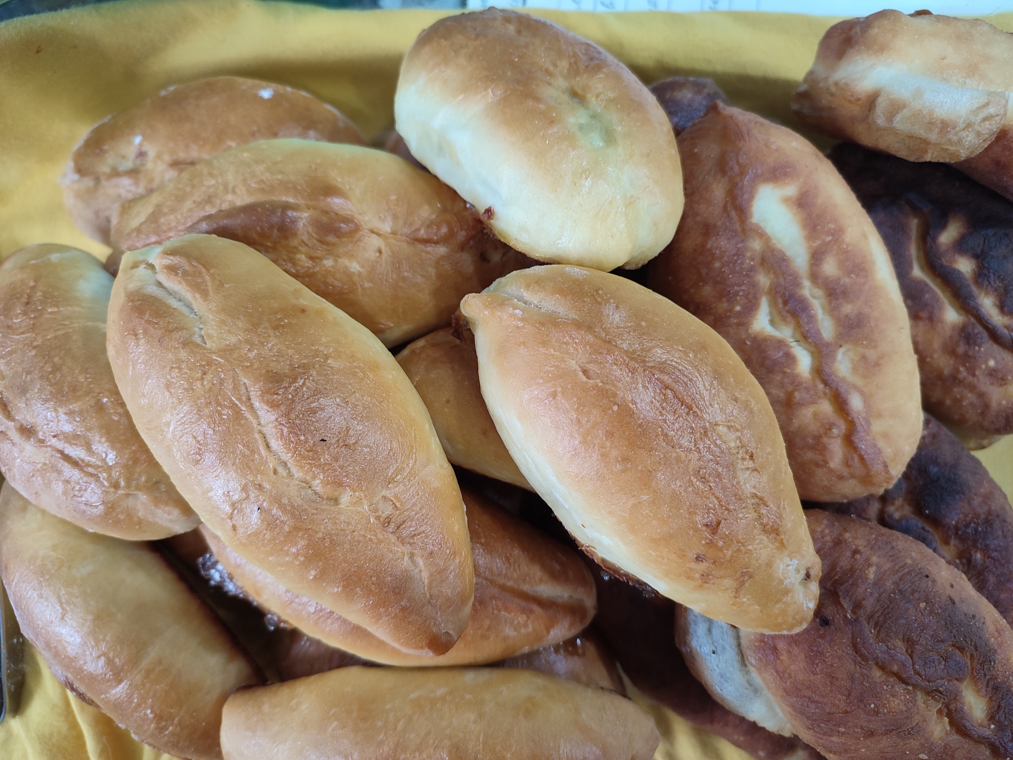 A pile of baked goods, possibly pirozhki or small bread rolls, are arranged on a yellow cloth. The baked goods are oval-shaped and have a golden-brown crust. Some have a lighter, almost white, appearance, while others are darker and appear to be more thoroughly baked, with some areas exhibiting a burnt or caramelized look. Several of the items have a visible slit or incision on their surface. The lighting suggests it could be daytime, but there are no other external cues to determine the exact time of day or weather. The setting appears to be indoors, given the uniform lighting and the presence of the cloth as a tablecloth or serving surface. No people are visible in the image, and no text is present. The overall impression is of a food item ready for consumption, possibly at a bakery, market stall, or during a gathering.