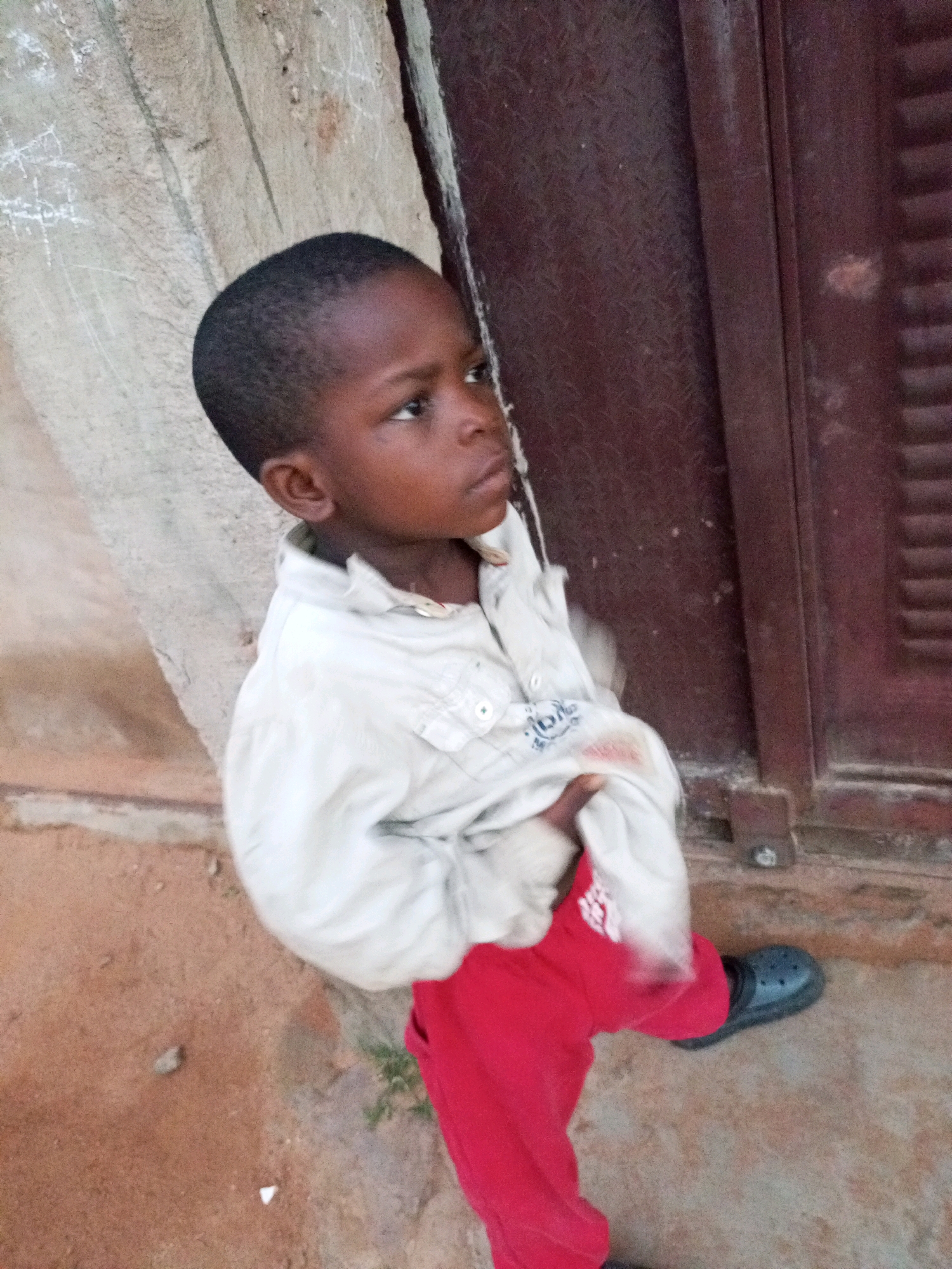 A young Black male child, appearing to be approximately 5 years old, stands outdoors in what is likely a residential area in Tempure, Nigeria. The child is wearing a light-colored, collared shirt with buttons down the front and bright red trousers. A dark-colored clog-style shoe is visible on the child's right foot. The child is looking off to the right of the frame with a slightly downcast gaze. The background consists of a rough, textured concrete wall on the left, and a dark brown metal door or gate on the right, which has horizontal slats and a visible latch. The ground beneath the child is a reddish-brown, sandy or dusty surface, with some small pebbles and debris visible. A faint hint of green vegetation can be seen in the lower right portion of the image. The lighting suggests it is daytime, with soft shadows.