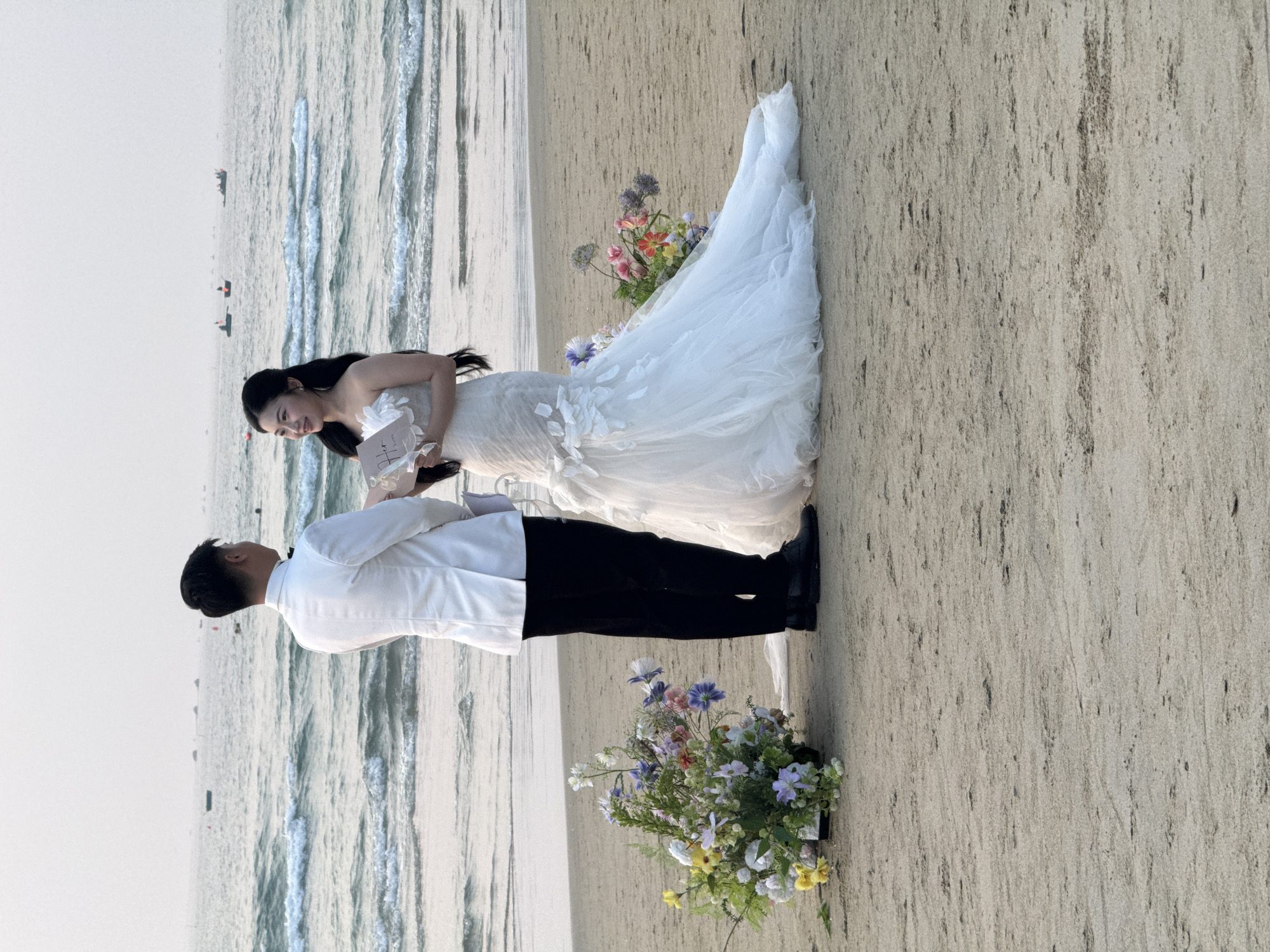 A wedding is occurring on a beach in what appears to be Đà Nẵng, Vietnam. The bride, in a strapless white gown with floral appliques, stands with the groom, dressed in a white shirt and black pants. They are positioned on the sand with the ocean in the background. A bouquet of colorful flowers sits on the sand to one side of the couple, and another sits nearby. The bride looks down at the groom, appearing to be reading from a small white booklet.