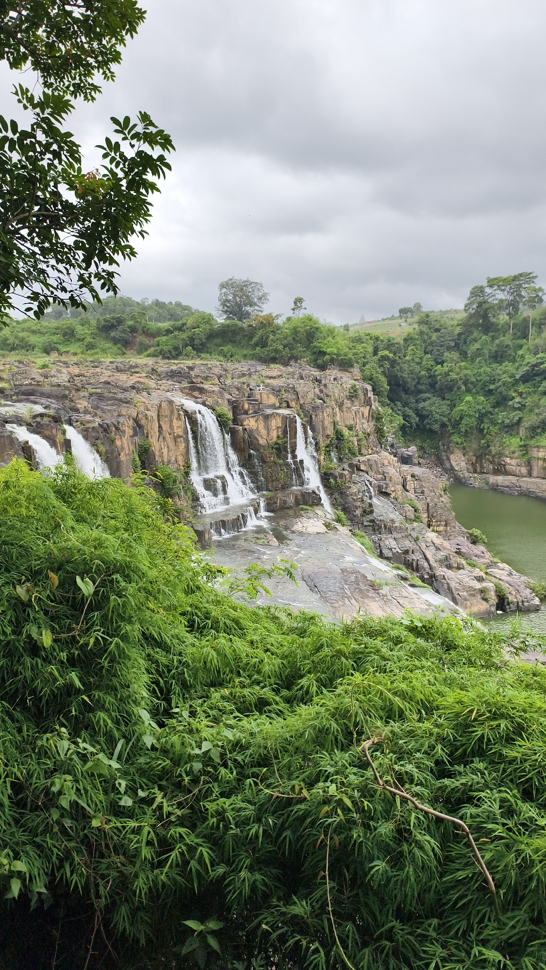This is a landscape photograph of a tiered waterfall in a natural setting. The waterfall cascades down a series of rocky ledges, with water flowing into a calm body of water at the base. Lush green vegetation, including trees and bamboo, surrounds the waterfall and fills the foreground. The sky is overcast with gray clouds. The scene appears to be located in the vicinity of Tân Phú, Vietnam, given the prevalence of tropical vegetation. There are no people or animals visible in the frame. The overall impression is one of a serene and untouched natural environment.