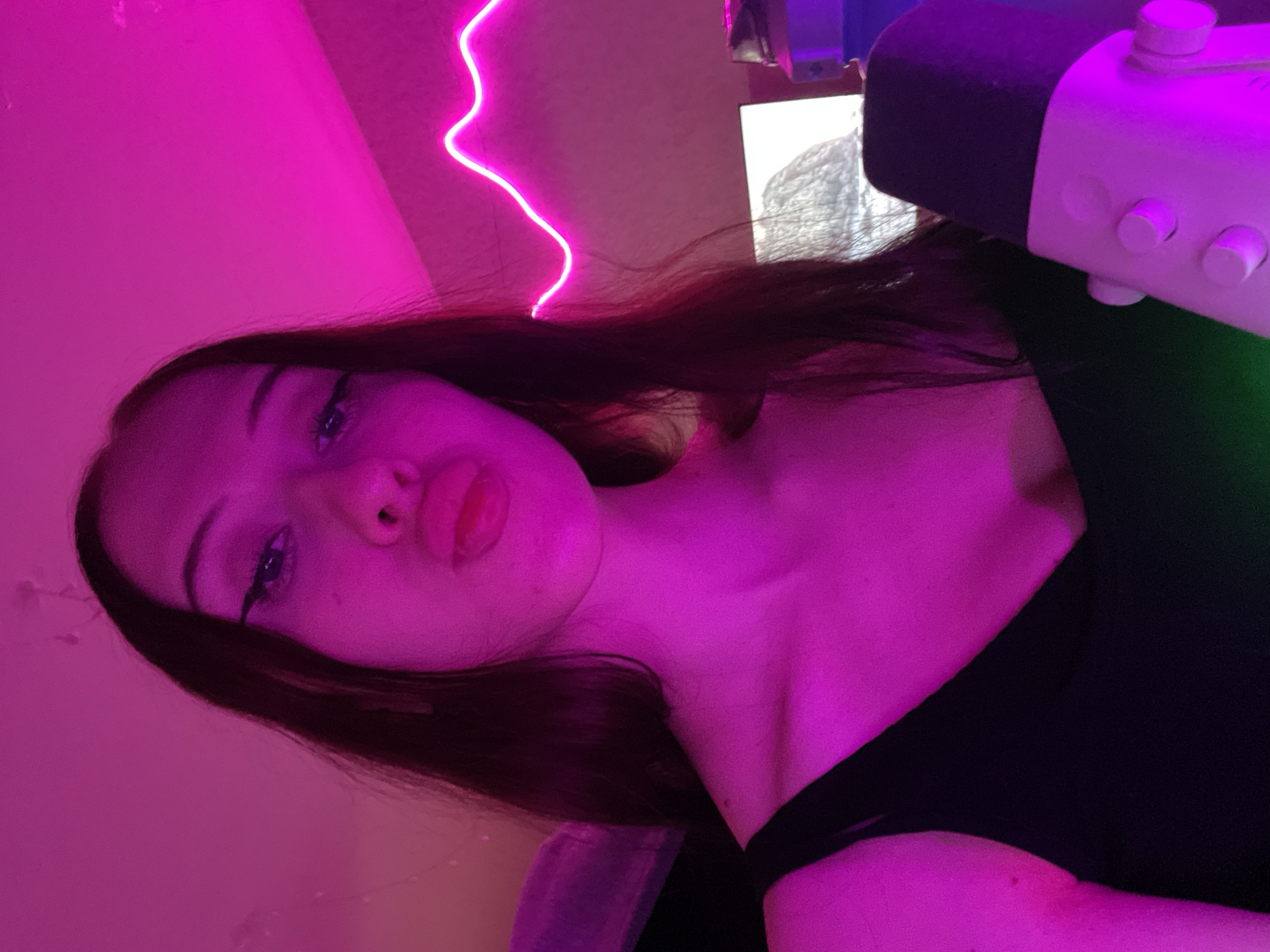The image is a close-up selfie of a young woman with long, dark hair and winged eyeliner. She is wearing a black top and her face is illuminated by bright pink light, possibly from a neon sign or LED lighting, which casts a purple hue on her skin. She is making a pouty face and holding a white microphone with black accents in her right hand. In the background, a computer monitor is visible, displaying an image of what appears to be a snowy landscape or a textured surface. A pink neon light strip is also visible on the wall behind her, forming a wave-like pattern. The overall environment suggests a room, possibly a home studio or gaming setup, given the presence of the microphone and computer. There is no indication of the location being in Votkinsk, Russia.