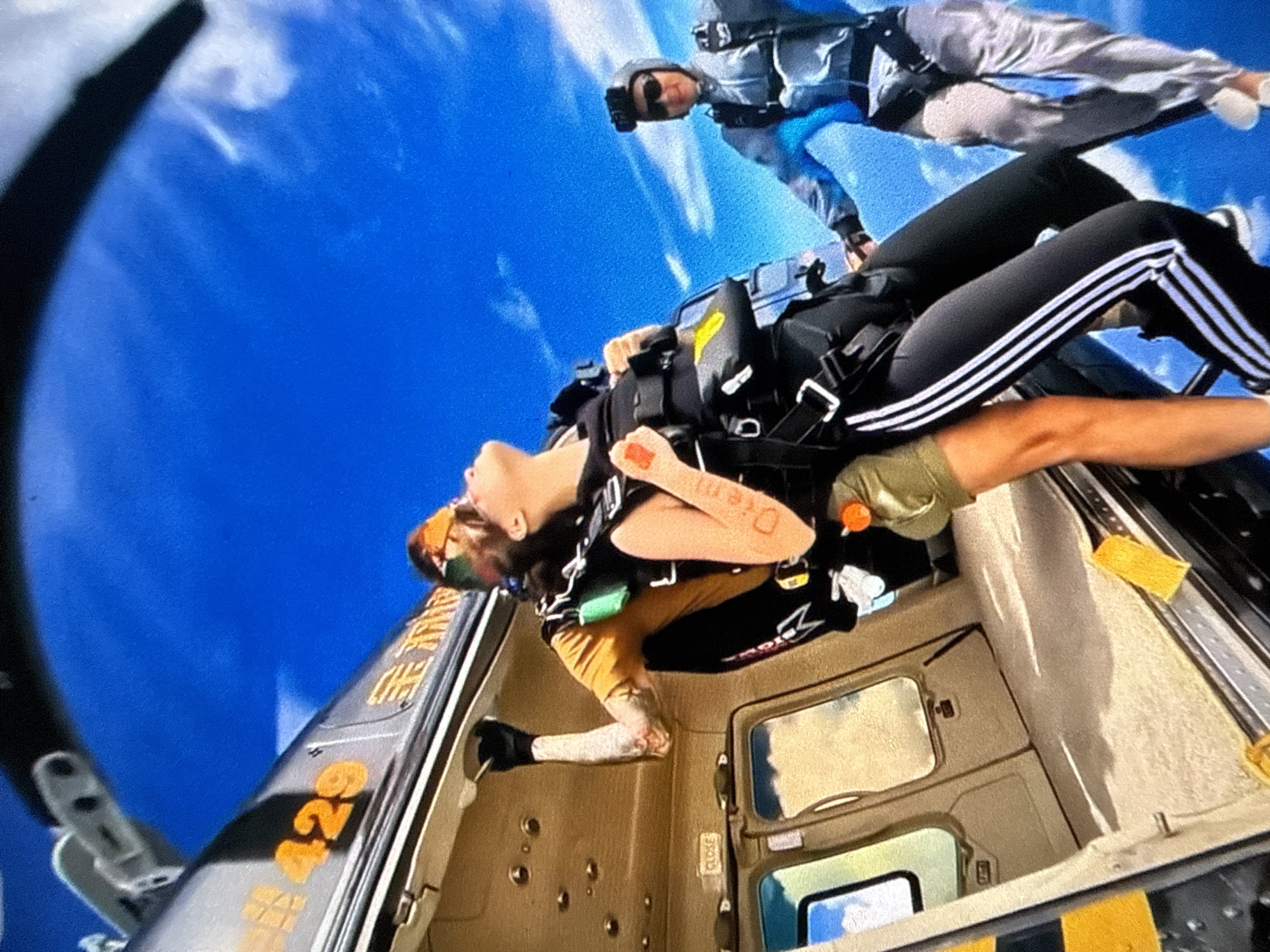 The image depicts two individuals engaged in skydiving, seemingly captured from the perspective of a helicopter. One person is in close proximity to the helicopter, preparing to jump, while the other is already in freefall above. The skydiver near the helicopter is equipped with a parachute harness and wearing black pants with white stripes down the side. They have writing on their arm. The other skydiver is wearing a grey jumpsuit and helmet. The interior of the helicopter is visible, displaying the door and various control elements. The number "429" is visible on the side of the helicopter. The environment features a bright blue sky dotted with a few clouds. The location is unknown.