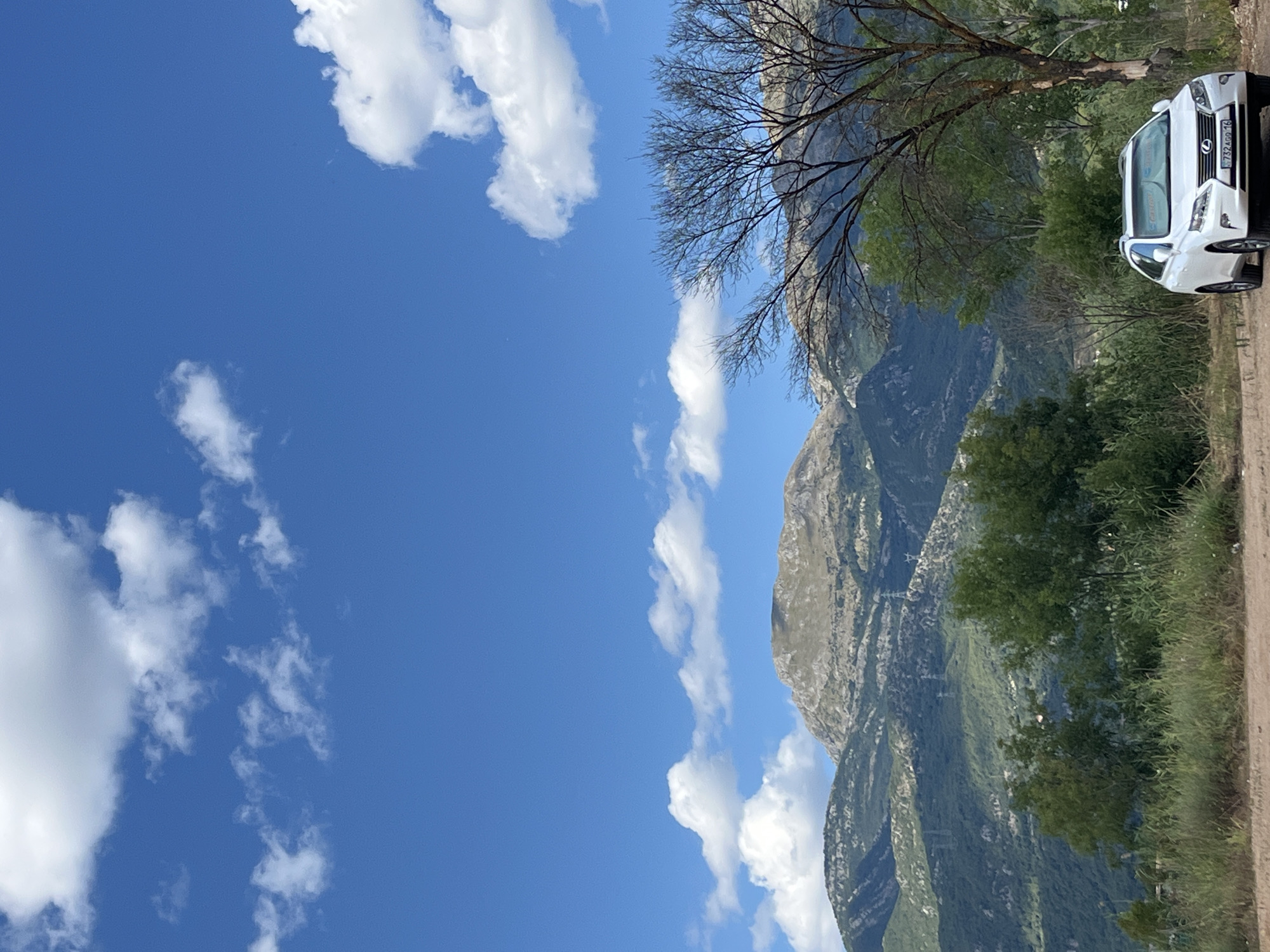 The image depicts a white Lexus SUV parked on a dirt road with a mountainous landscape in the background. The sky is a clear blue with scattered white clouds. The SUV is positioned in the lower right quadrant of the image, facing slightly to the left. The foreground consists of dry grass and dirt. Behind the car, there are several trees, some with sparse foliage and one larger tree with bare branches in the foreground. The middle ground is filled with lush green trees and vegetation, leading up to the rugged, green mountains in the background. The mountains exhibit layered formations and appear to be sparsely populated. The scene suggests an outdoor setting, possibly a scenic route or a viewpoint near Prijevor, Montenegro, given the context. There are no people visible in the image. The lighting suggests it is daytime, with sunlight casting shadows.