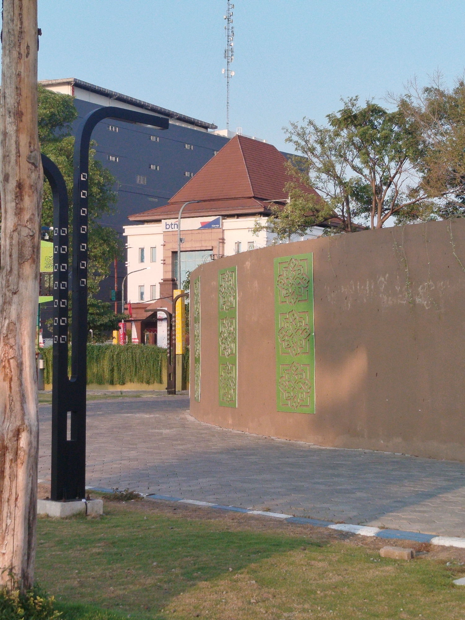 The image captures a street scene in Sidoarjo, Indonesia, on a clear day with a pale blue sky. In the foreground, a textured tree trunk stands to the left, with a black metal post adorned with square cutouts to its right, planted in a concrete base. Behind this, a paved pathway with interlocking square stones leads towards the background. To the right of the pathway is a curved, light brown wall with green decorative panels featuring intricate patterns.
In the mid-ground, a modern dark blue building with small rectangular windows is visible, partially obscured by trees. To its right, a building with a reddish-brown tiled roof and white walls stands, featuring a sign with "btn" and a blue logo. Further back, a telecommunications tower with multiple antennas rises against the sky. Lush green hedges line the area behind the paved pathway, separating it from the buildings. The overall environment suggests a well-maintained urban or semi-urban area. There are no people visible in the image.
