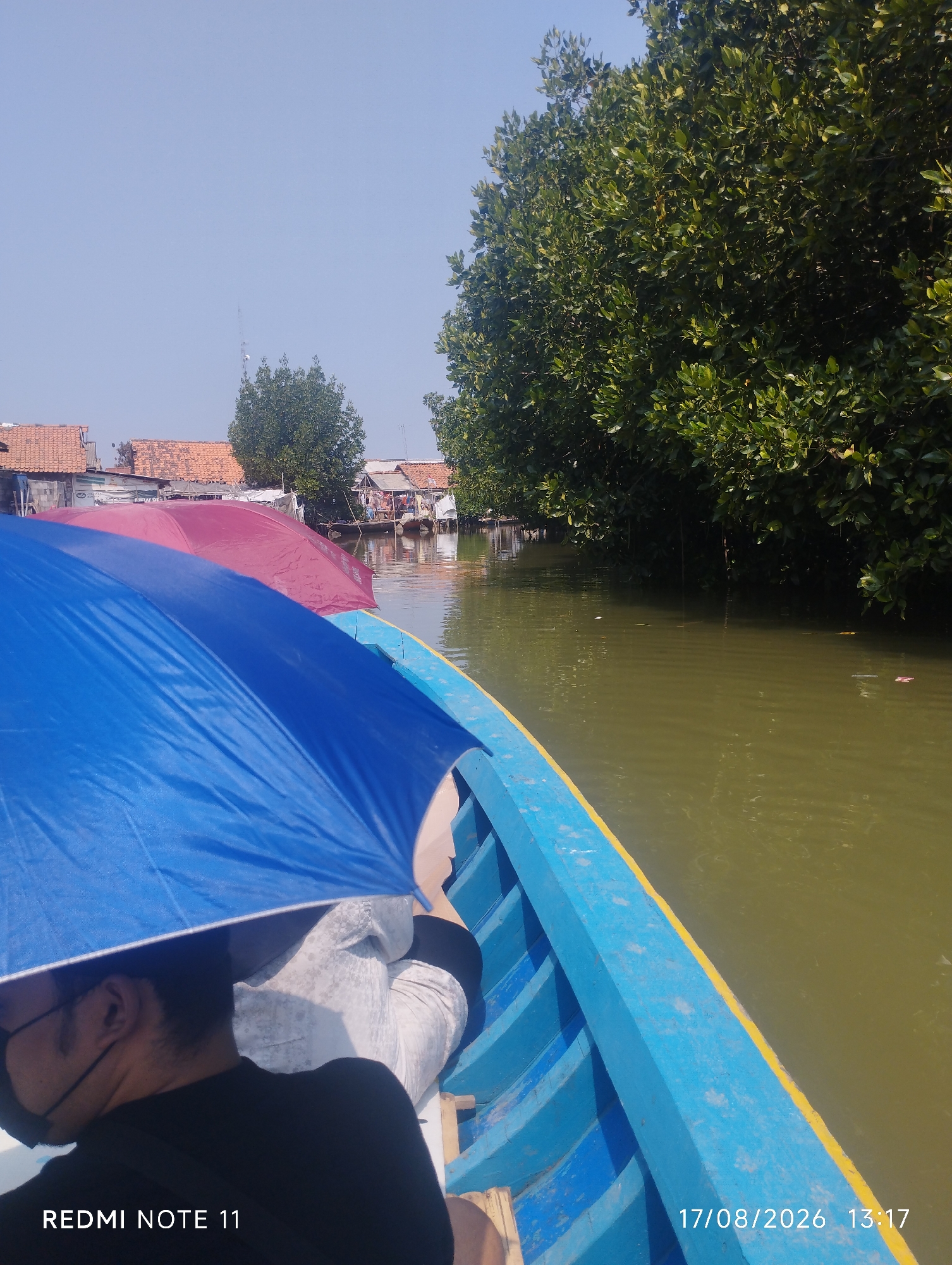 This is a photograph taken from a boat on a river, likely in a mangrove area. The boat, painted bright blue with a yellow trim, carries at least two people. In the foreground, a person wearing a black shirt and a face mask is partially visible, holding a blue umbrella. Another umbrella, maroon in color, is seen further down the boat. The river water is murky green. On the right side of the river, dense green mangrove trees line the bank. On the left side of the river, several stilt houses with red tile roofs are visible, suggesting a settlement. The sky is clear and blue. A date and time stamp at the bottom right corner reads "17/08/2026 13:17", and a watermark at the bottom left corner says "REDMI NOTE 11". While the prompt requests the location in Karawang, Indonesia, this specific location cannot be definitively determined from the image alone.