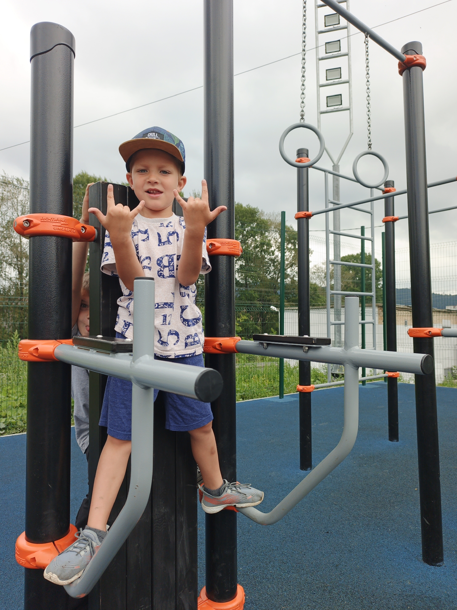 A young boy, wearing a cap, a patterned t-shirt, and blue shorts, stands on a metal bar of an outdoor exercise apparatus. He is making a "rock on" gesture with both hands. Another child, partially visible, is peeking from behind a black pole of the apparatus. The setting appears to be a public playground in Zlatoust, Russia, with a blue rubberized surface under the equipment and a chain-link fence in the background. The sky is overcast.