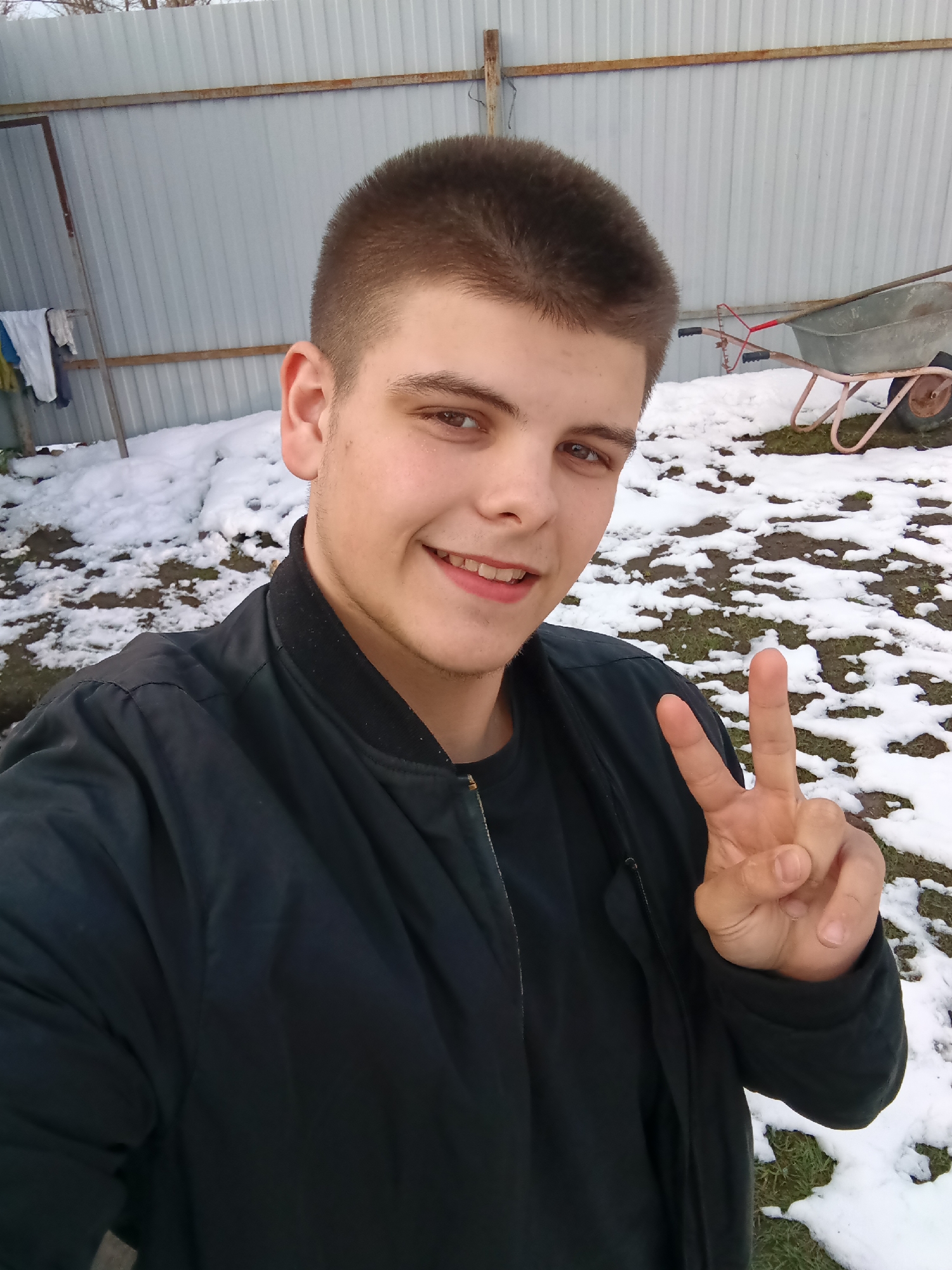 A young man is taking a selfie outdoors in what appears to be a snowy area. He is wearing a black jacket and making a peace sign with his right hand. The background shows a metal fence, patches of snow, and a wheelbarrow. Based on the city mentioned, Andreyevo, Russia, the environment suggests a rural or suburban area.