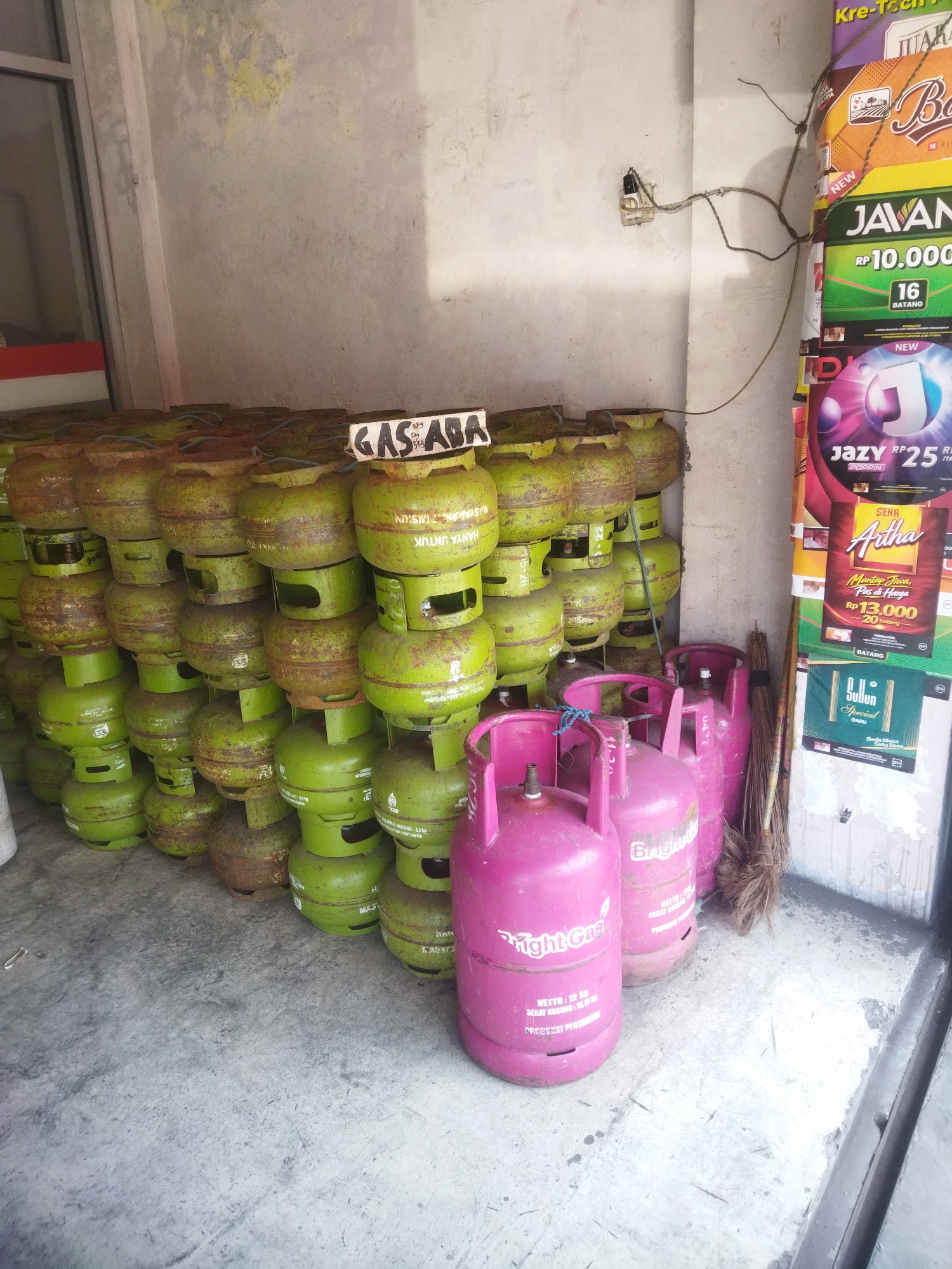 The image shows a storage area with numerous gas cylinders stacked high. In the foreground, there are several bright pink gas cylinders, some with "Bright Gas" branding visible. Behind these, a large quantity of green gas cylinders are stacked in rows, some showing signs of rust. A sign with "GAS ABA" written on it is placed on top of the green cylinders. The environment appears to be an outdoor or semi-outdoor space with a concrete floor and a wall with peeling paint in the background. Advertising posters are visible on the right side of the image, suggesting a commercial setting. The overall scene depicts a retail or distribution point for gas cylinders in Giripurwo, Indonesia.