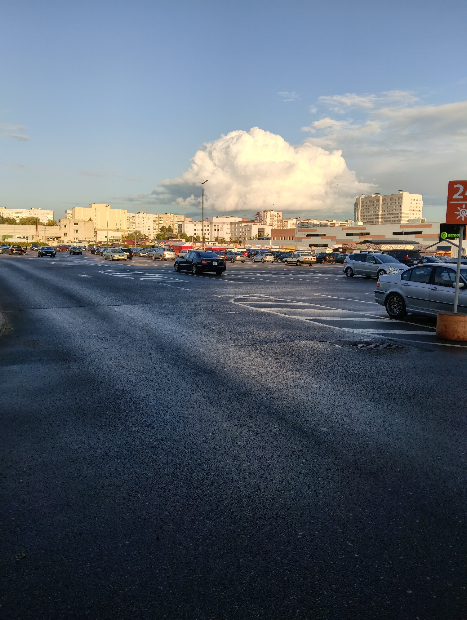 An outdoor, eye-level, long shot captures a parking lot filled with cars under a bright sky with a large, fluffy white cloud. The parking lot pavement is dark and wet, reflecting the sky and casting shadows from unseen objects on the left. Multiple cars are parked and driving in the lot, most of them silver or black. In the background, a cityscape of apartment buildings and commercial structures is visible. A red and white sign on the right side of the image shows the number "24" and a sun symbol. The lighting suggests it is late afternoon or early evening. This scene is likely in the city of Hrodna, Belarus, given the architectural style of the buildings and the overall urban environment.